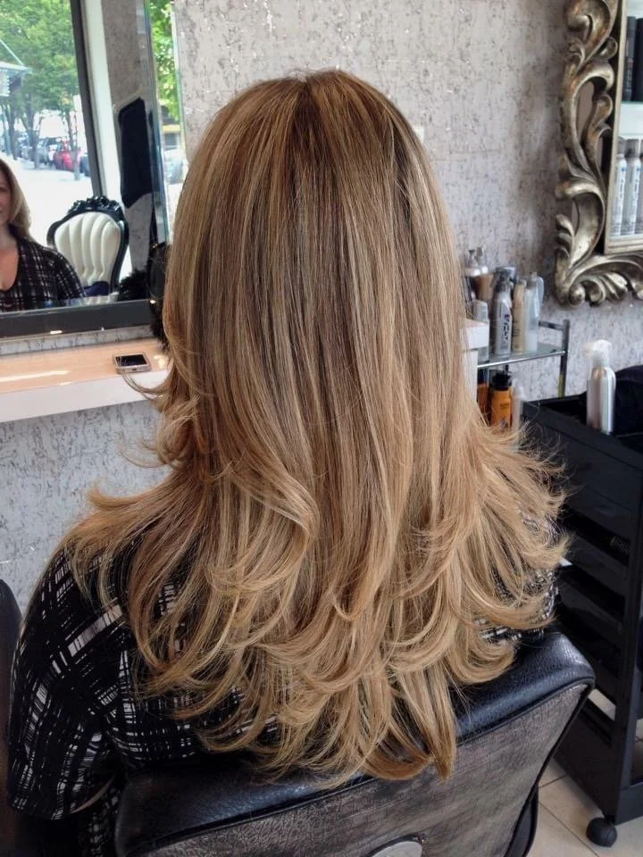 Back view of a woman with long wavy blonde hair sitting in a salon chair, with a mirror and salon products visible in the background.