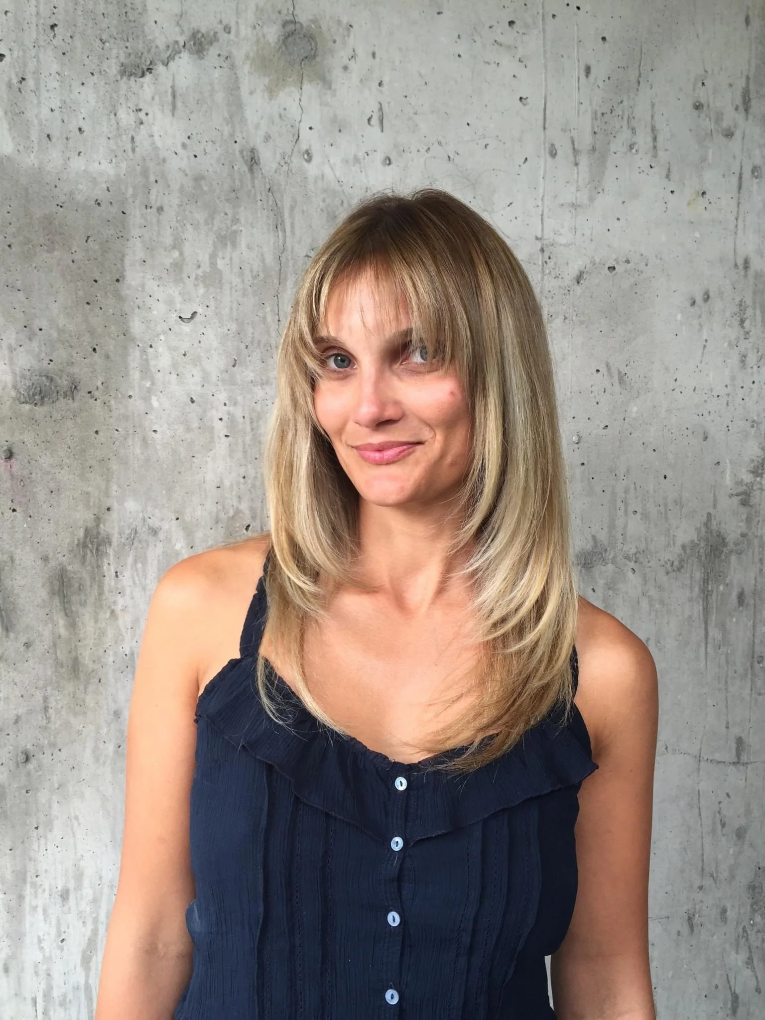 A woman with shoulder-length blonde hair, blue eyes, and a subtle smile, wearing a sleeveless navy blue top, standing against a textured gray concrete wall.