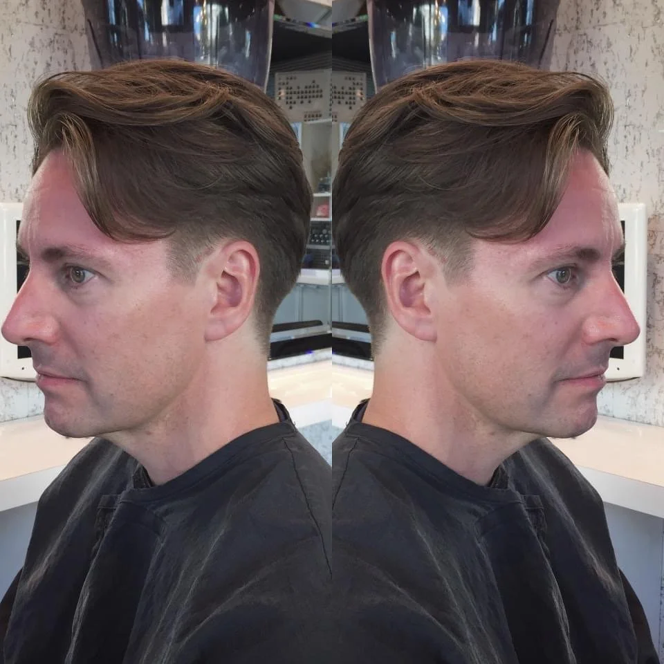 A mirrored image of a man with short brown hair, seated in a barbershop chair, looking to the right, with hair styled in a side-parted haircut.