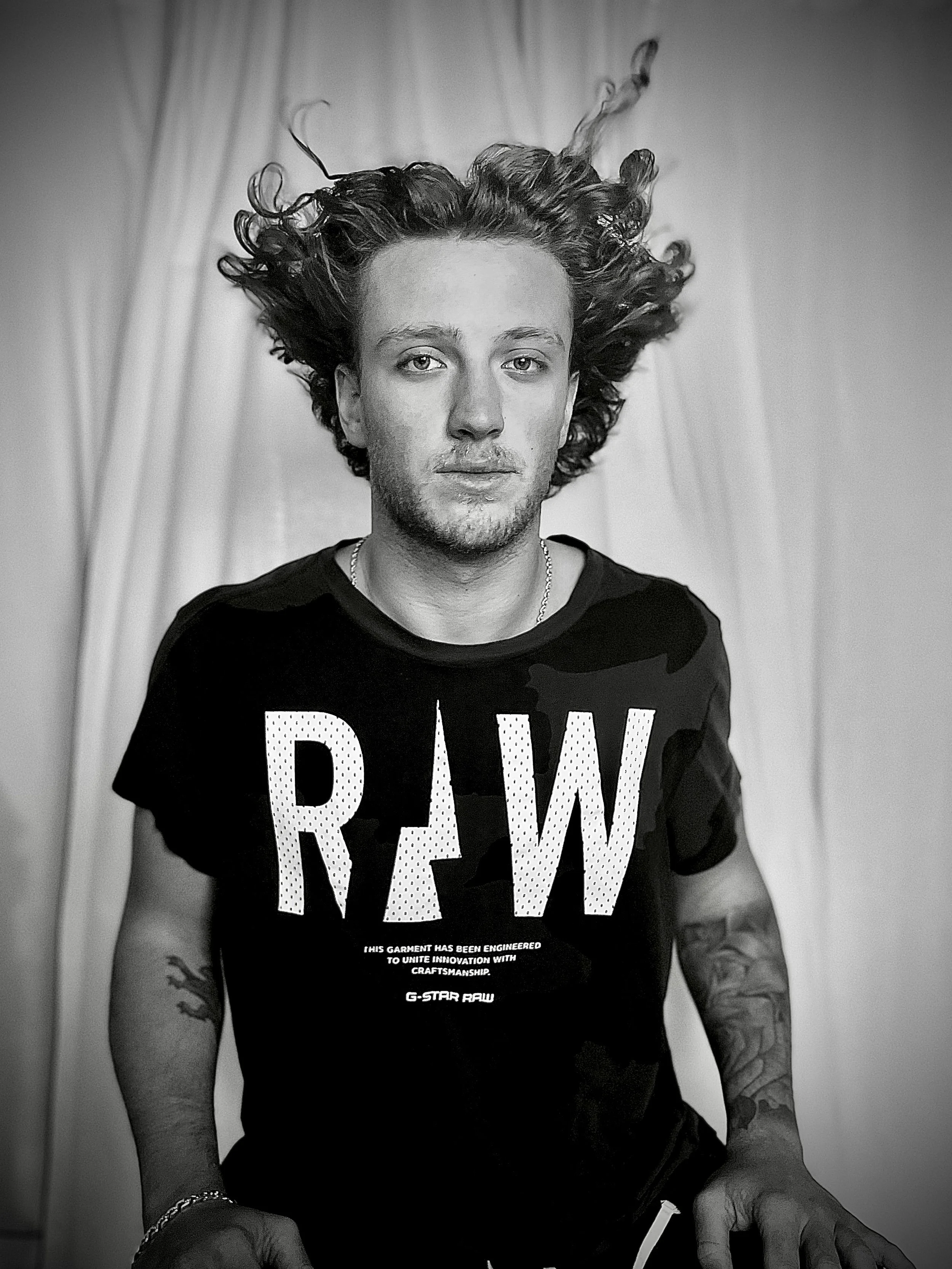 Person with curly hair wearing a t-shirt with 'RAW' text, black and white photo.