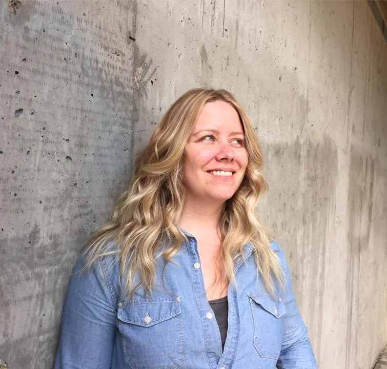 A woman with long blonde curly hair smiling and looking to her right, standing beside a concrete wall, wearing a light blue denim shirt.