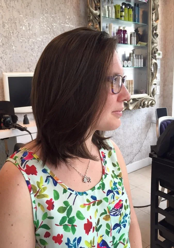 Profile of a woman with shoulder-length brown hair, glasses, wearing a colorful sleeveless floral top with birds, sitting in a salon with a mirror and various products in the background.