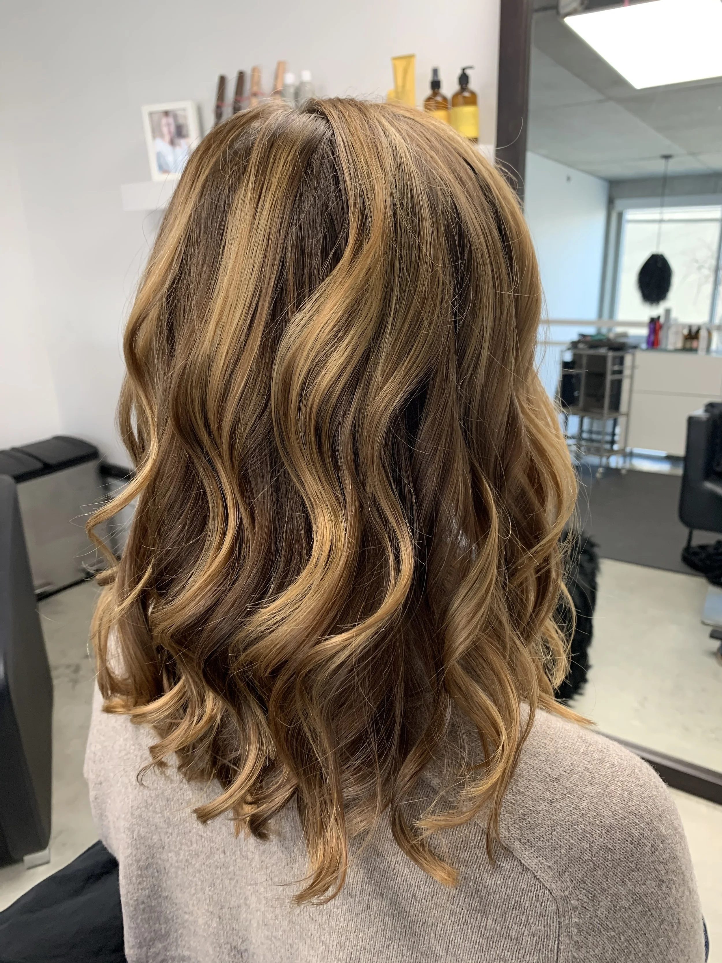 A woman with shoulder-length, wavy, light brown hair with highlights, sitting in a salon chair.