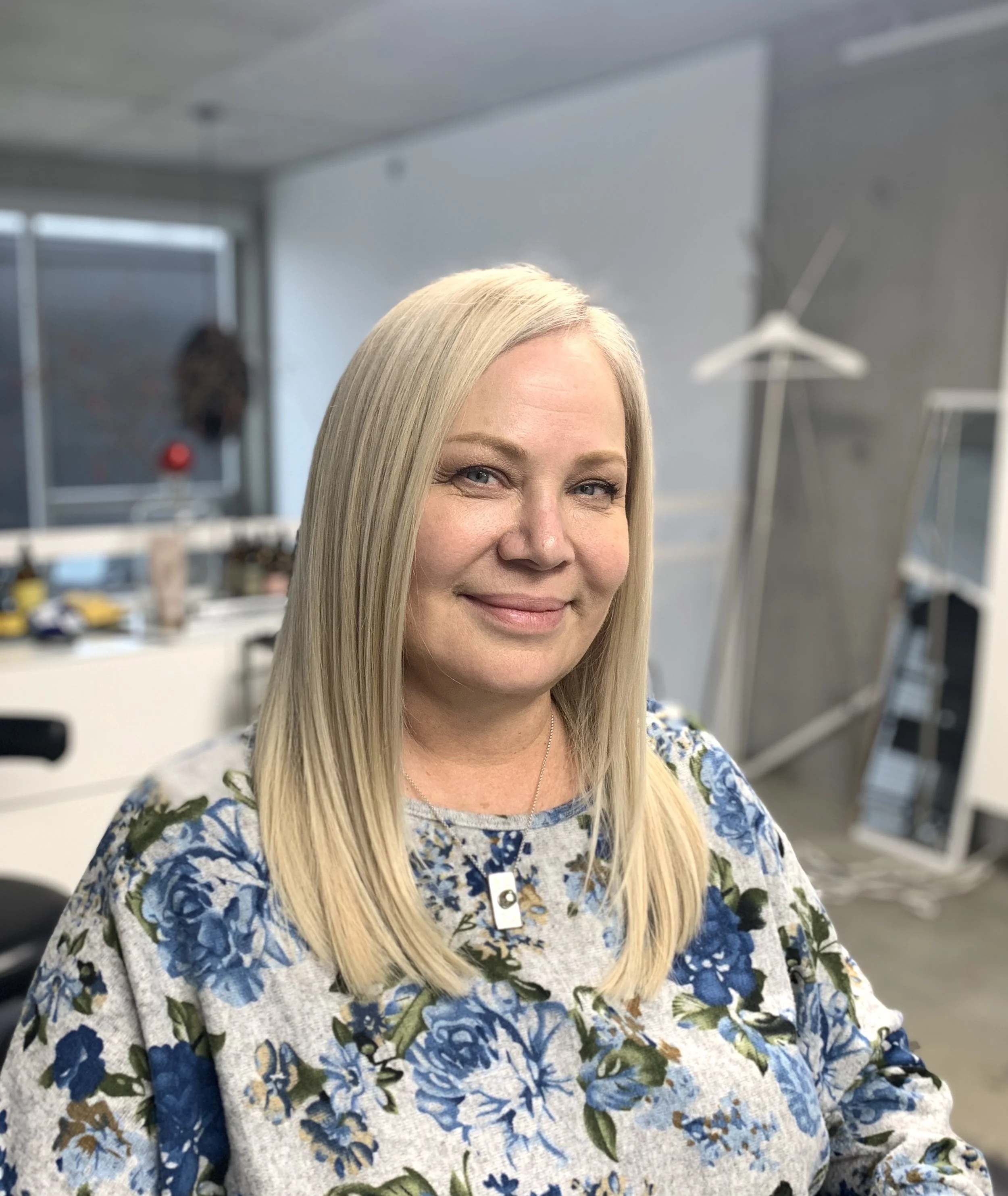 A woman with long blonde hair, wearing a floral sweater, smiling in an indoor office setting in front of a desk and blurred background.
