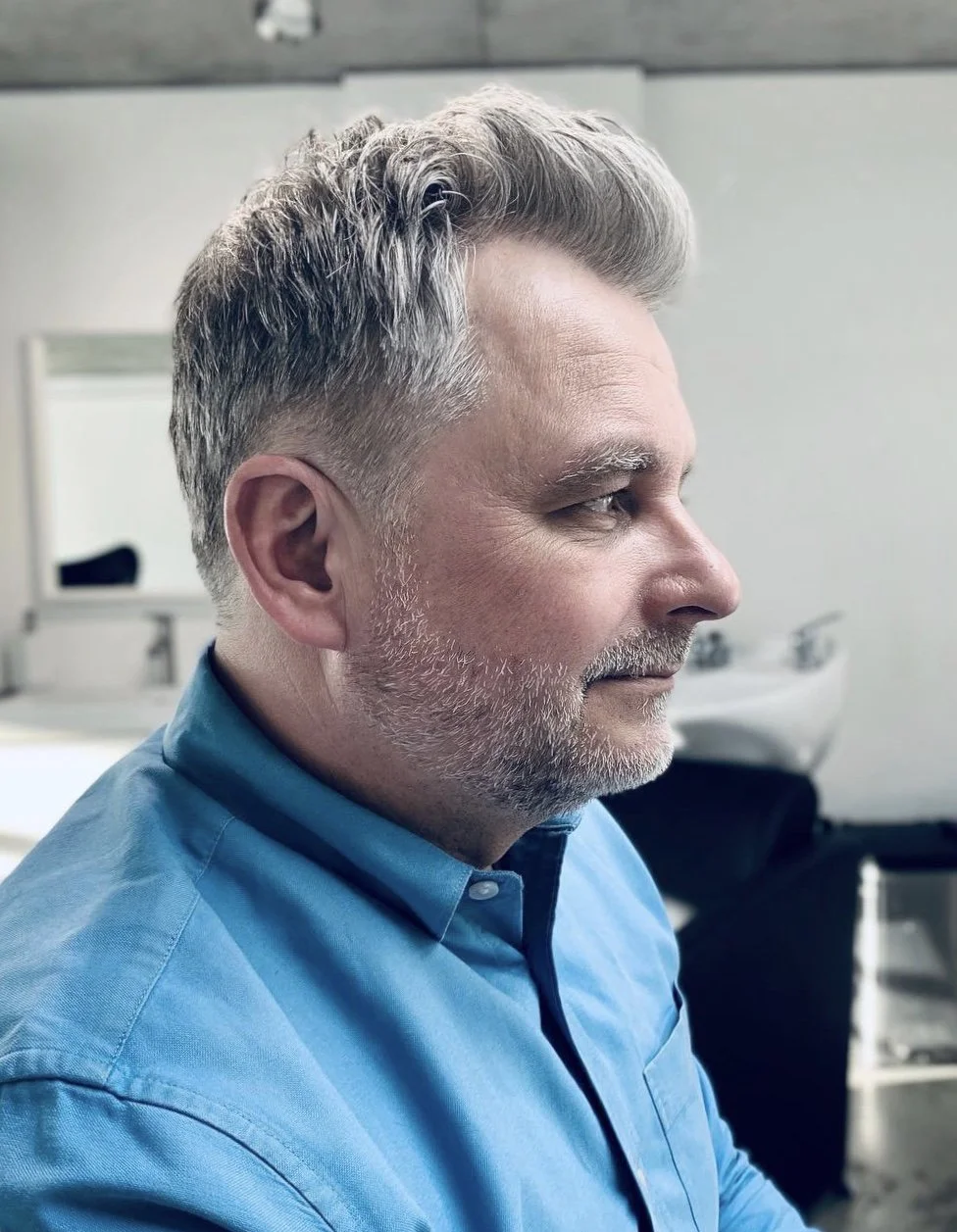 Man with gray hair and beard wearing a blue shirt sitting in a modern interior