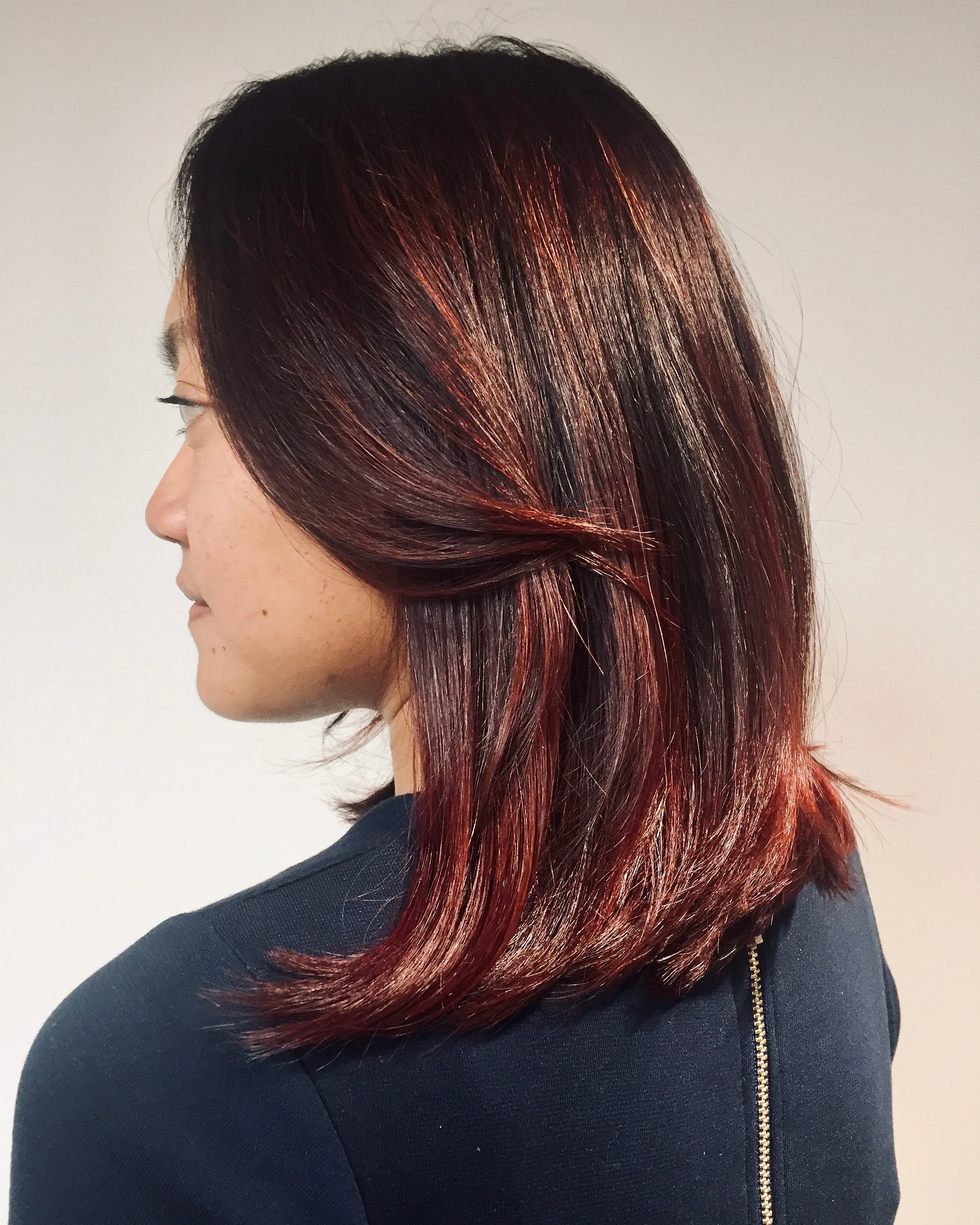 Side profile of a woman with shoulder-length, dark hair with red highlights, wearing a dark top with a gold zipper on the back.