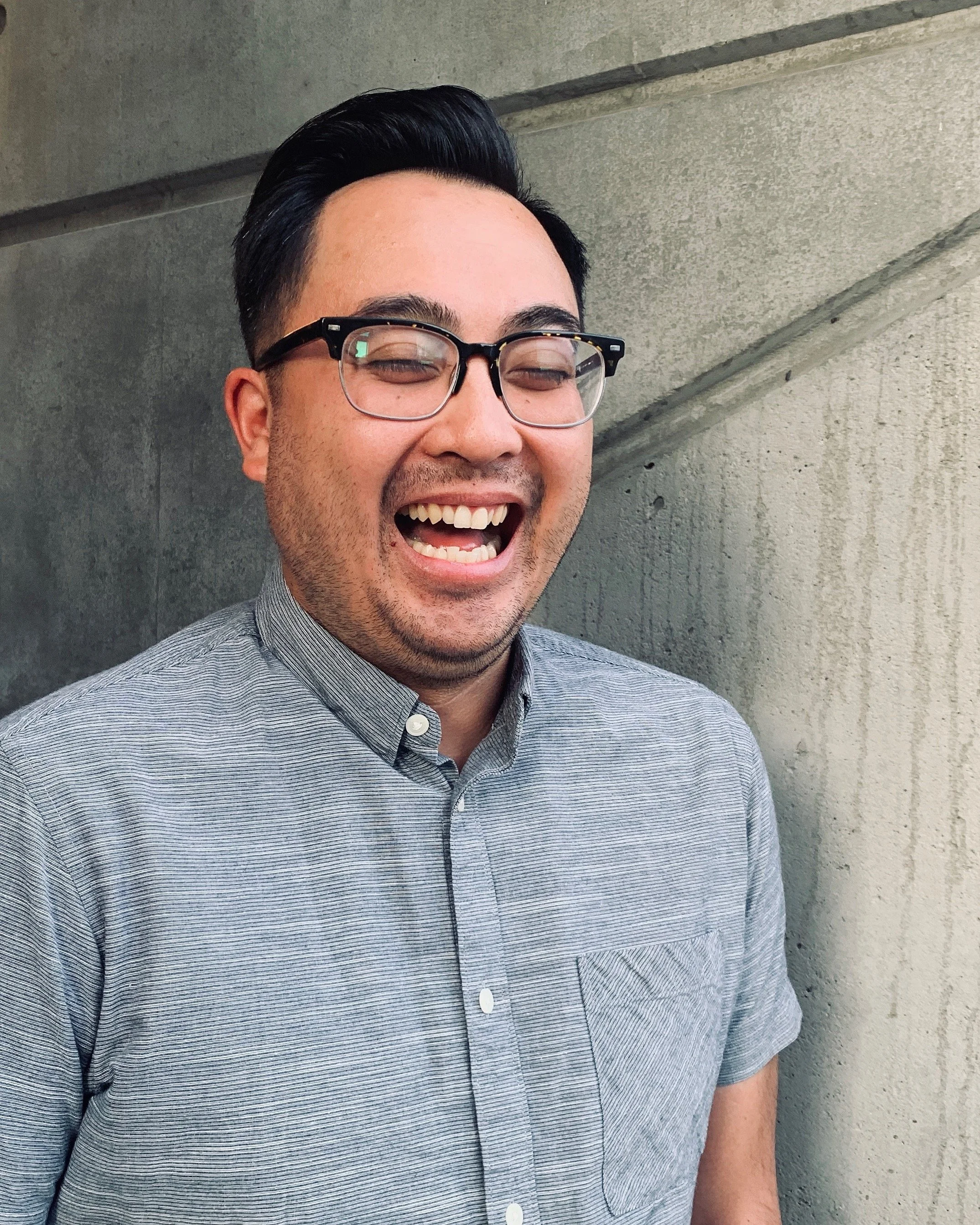 A young man with dark hair, glasses, and a gray button-up shirt is smiling and laughing against a gray concrete wall.