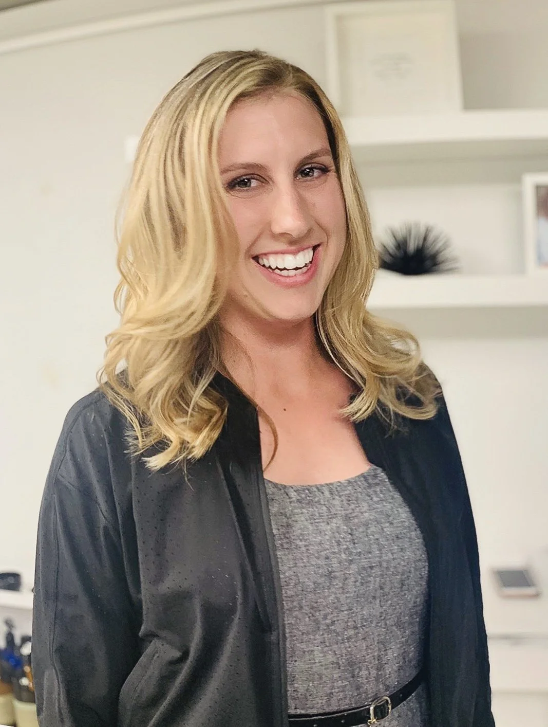 A smiling person with long blond hair wearing a black jacket over a gray top is standing indoors with white shelves in the background.