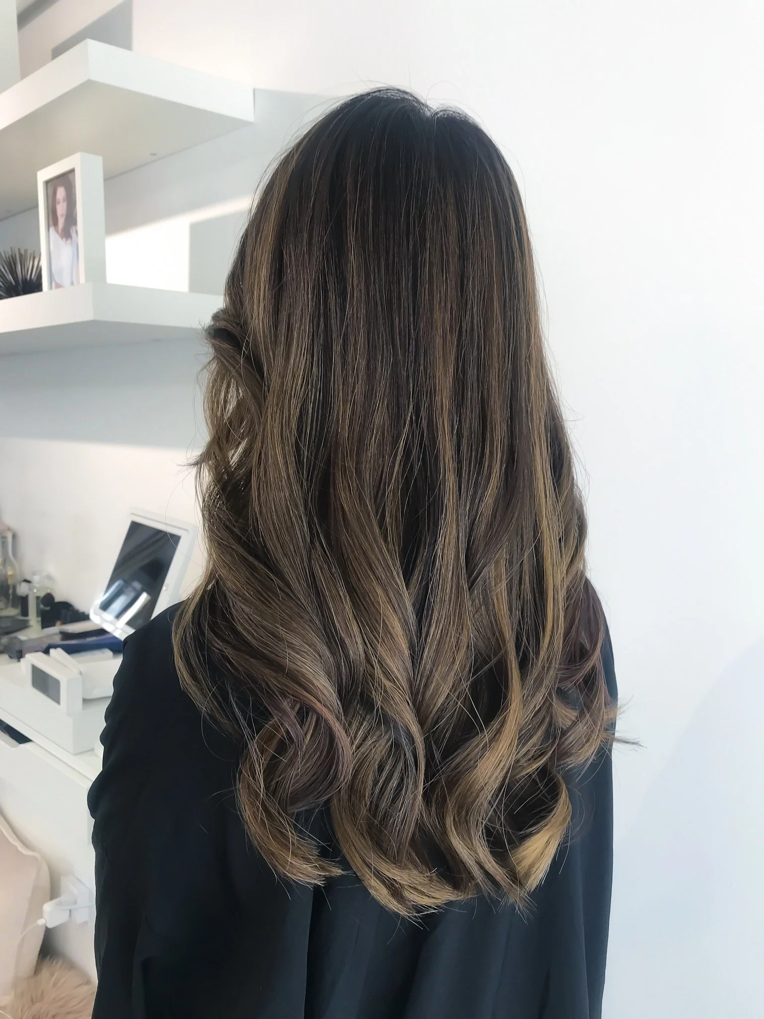 Back view of a person with long, wavy brown hair in a modern room with white shelves.