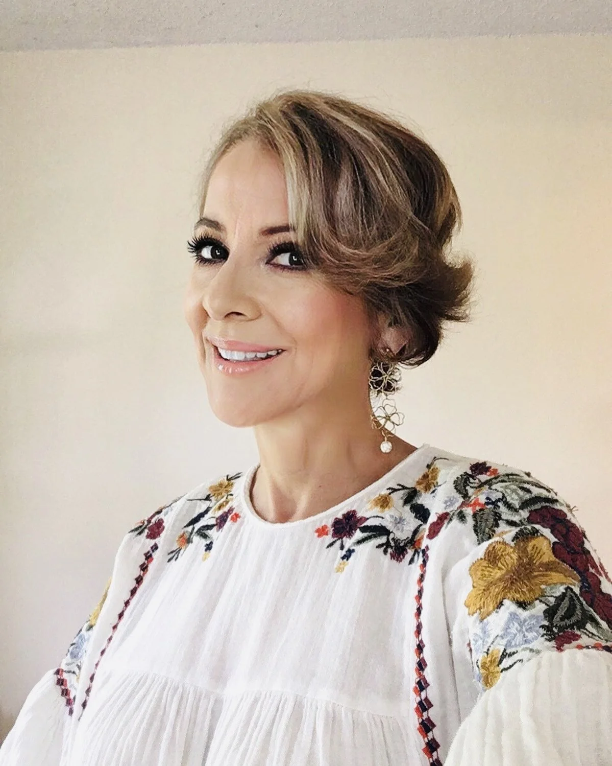 Smiling person wearing a white blouse with colorful floral embroidery, accessorized with earrings, against a neutral background.