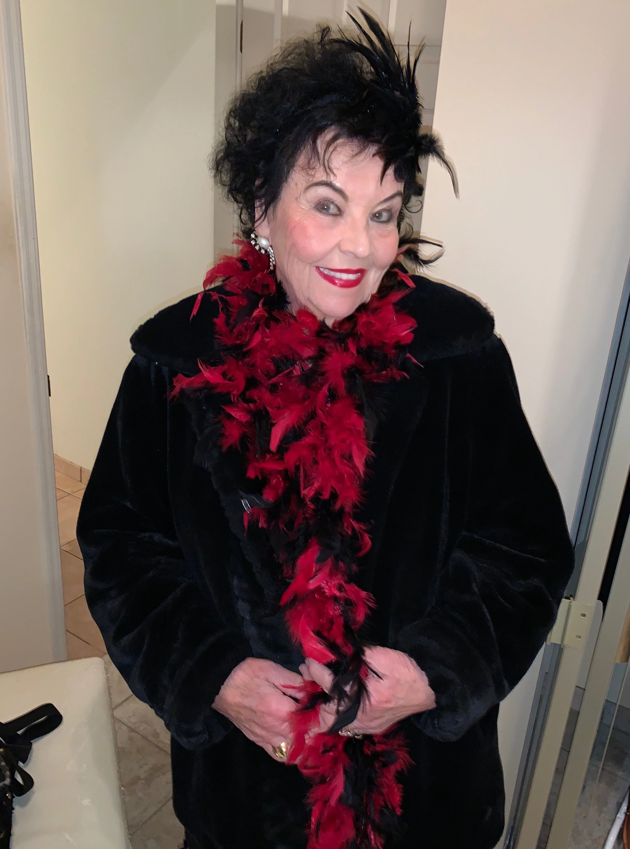 A woman with black curly hair wearing a black velvet coat and a red and black feathered scarf, smiling at the camera.