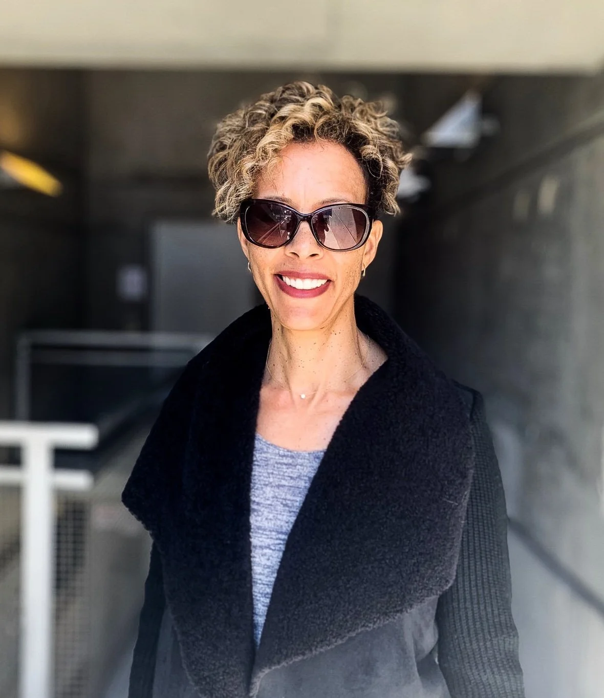 A woman with curly hair, wearing large sunglasses and a dark coat, smiling in a well-lit indoor setting.