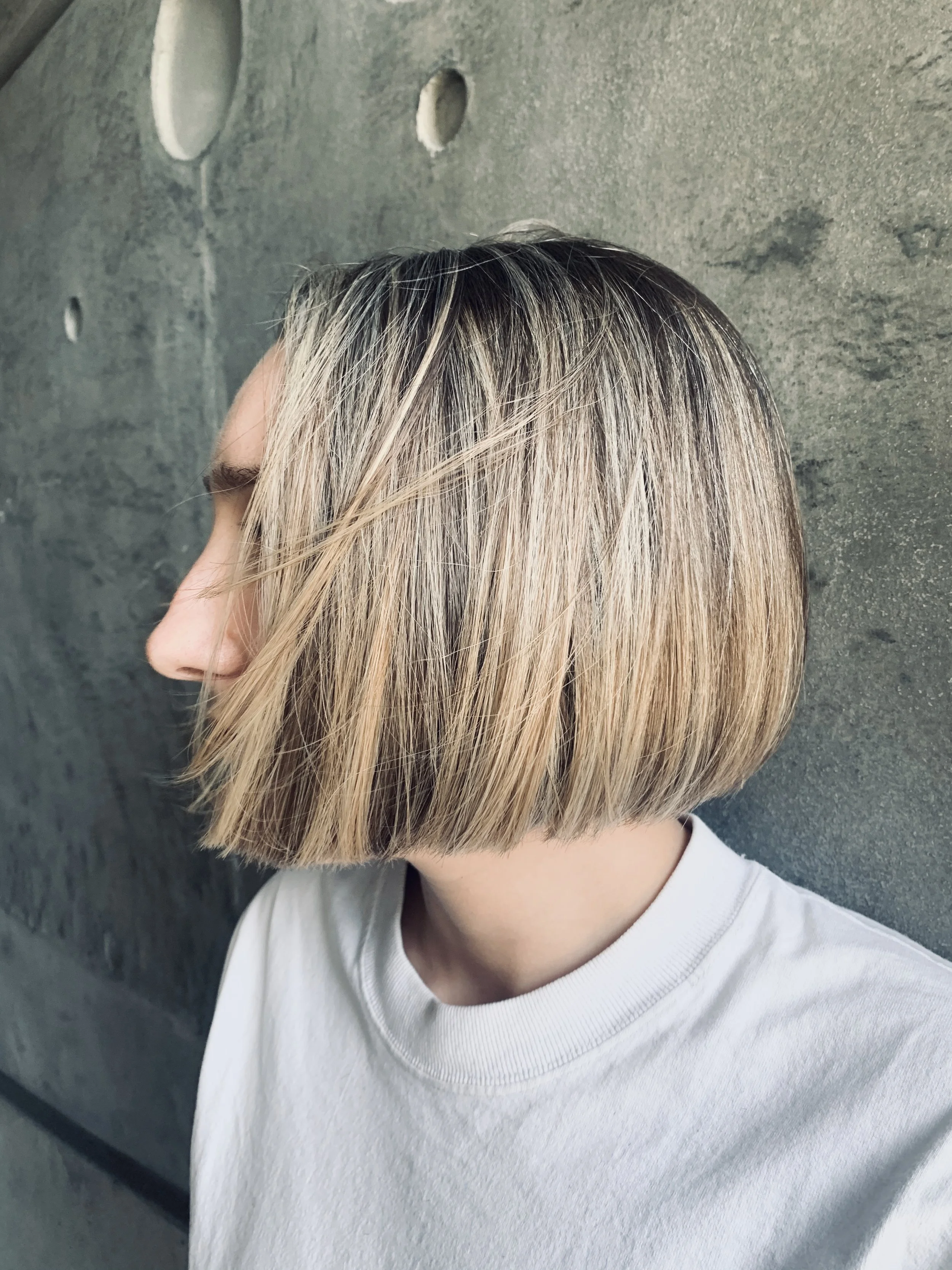 Side profile of a person with a blonde bob hairstyle, wearing a white shirt, standing against a textured concrete wall.