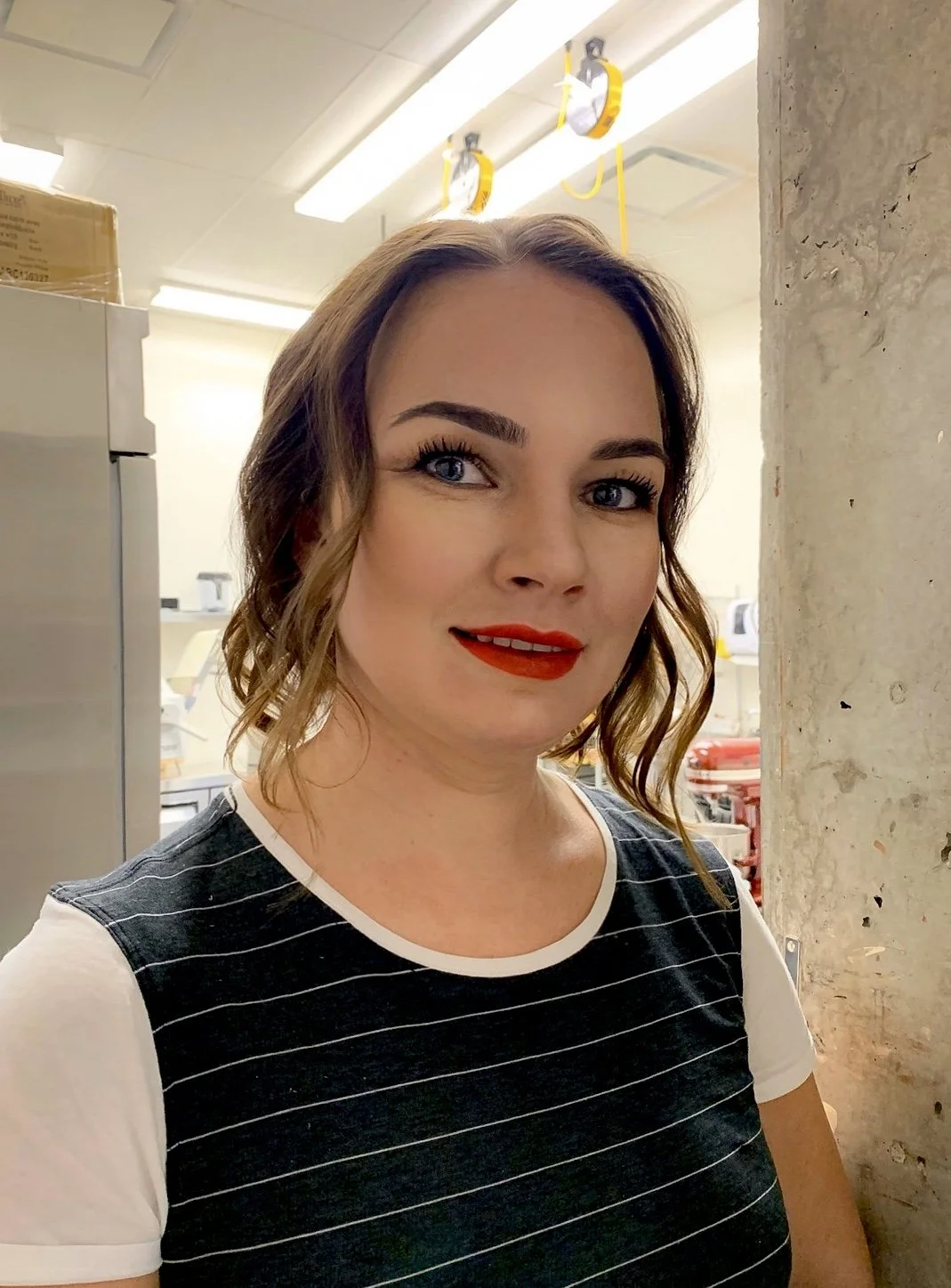A woman with wavy brown hair, red lipstick, and blue eyes wearing a black and white striped shirt with white sleeves, standing indoors near a concrete pillar with a background of kitchen equipment and equipment hanging from the ceiling.
