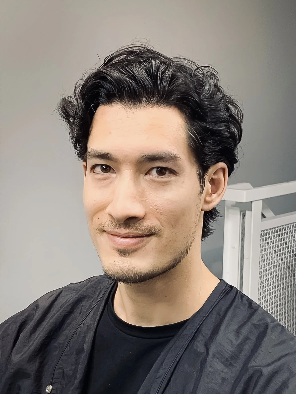 A man with dark, wavy hair and a slight beard, smiling at the camera, wearing a black shirt and a black cape, standing in front of a plain background with a metal mesh structure visible on the right.