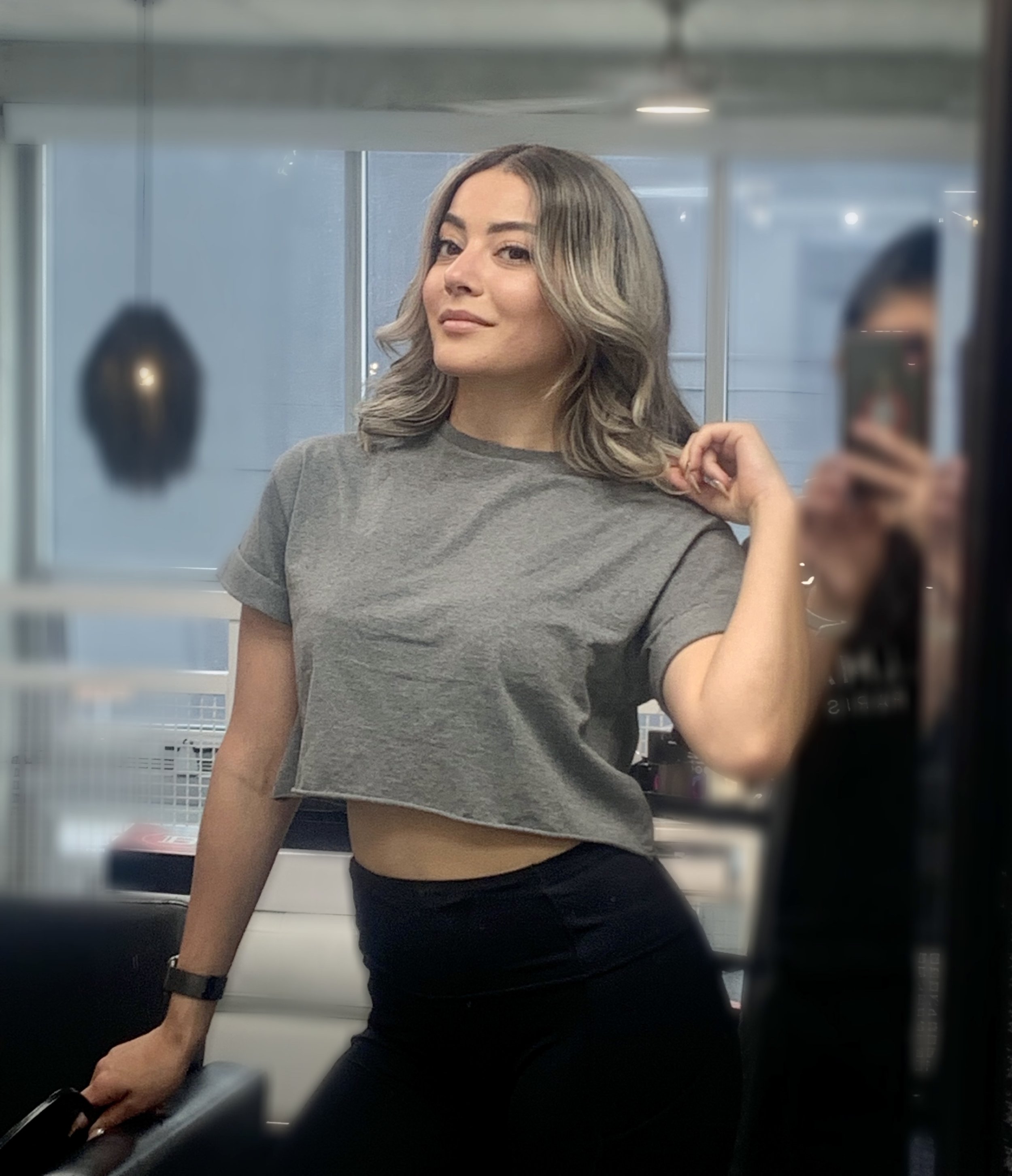 Woman in a gray crop top and black pants standing indoors, posing confidently with her hand on her hip, mirror reflection in the background.