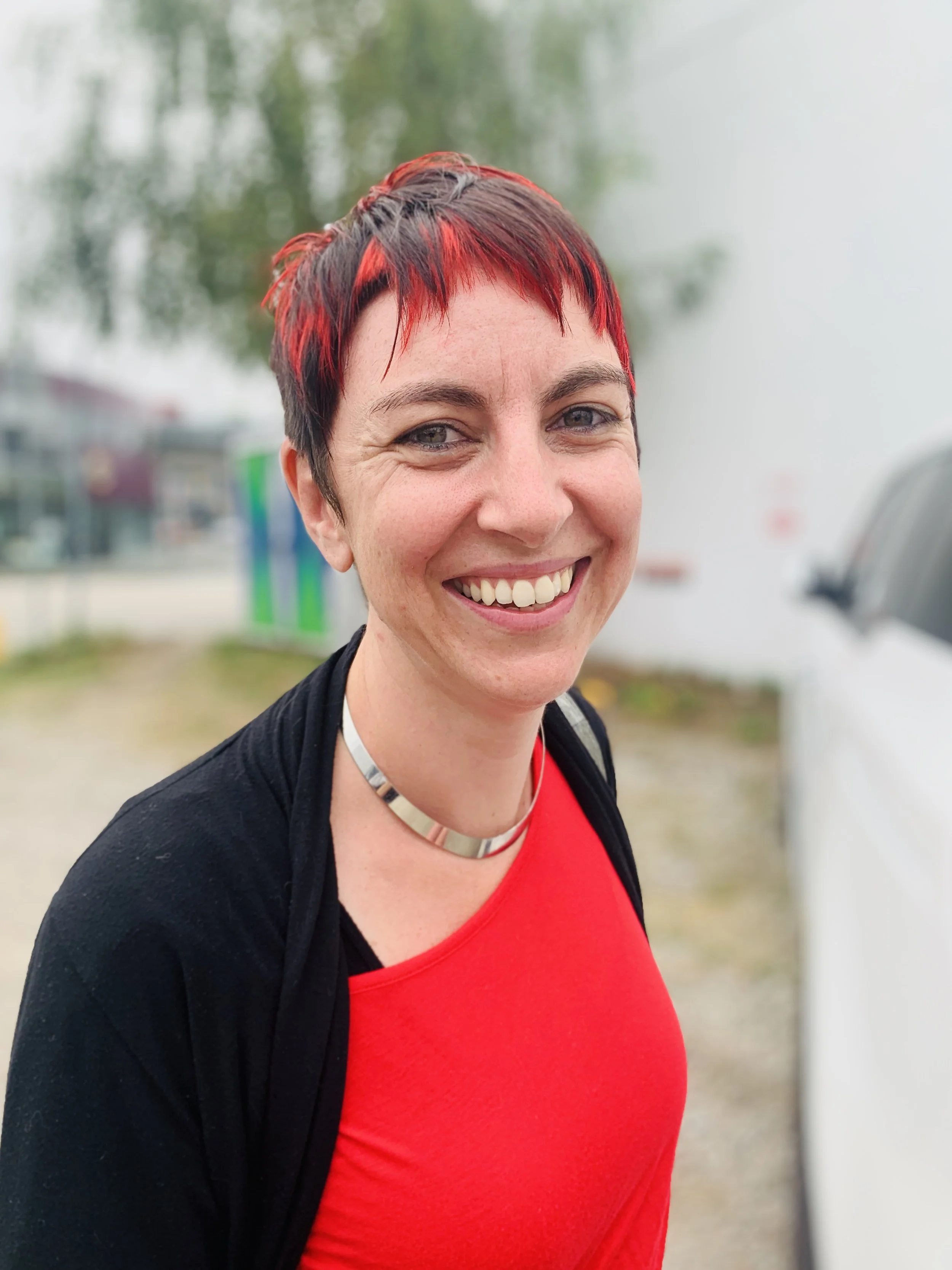 Smiling person with short red and black hair, wearing a red shirt and a black jacket, standing outdoors.