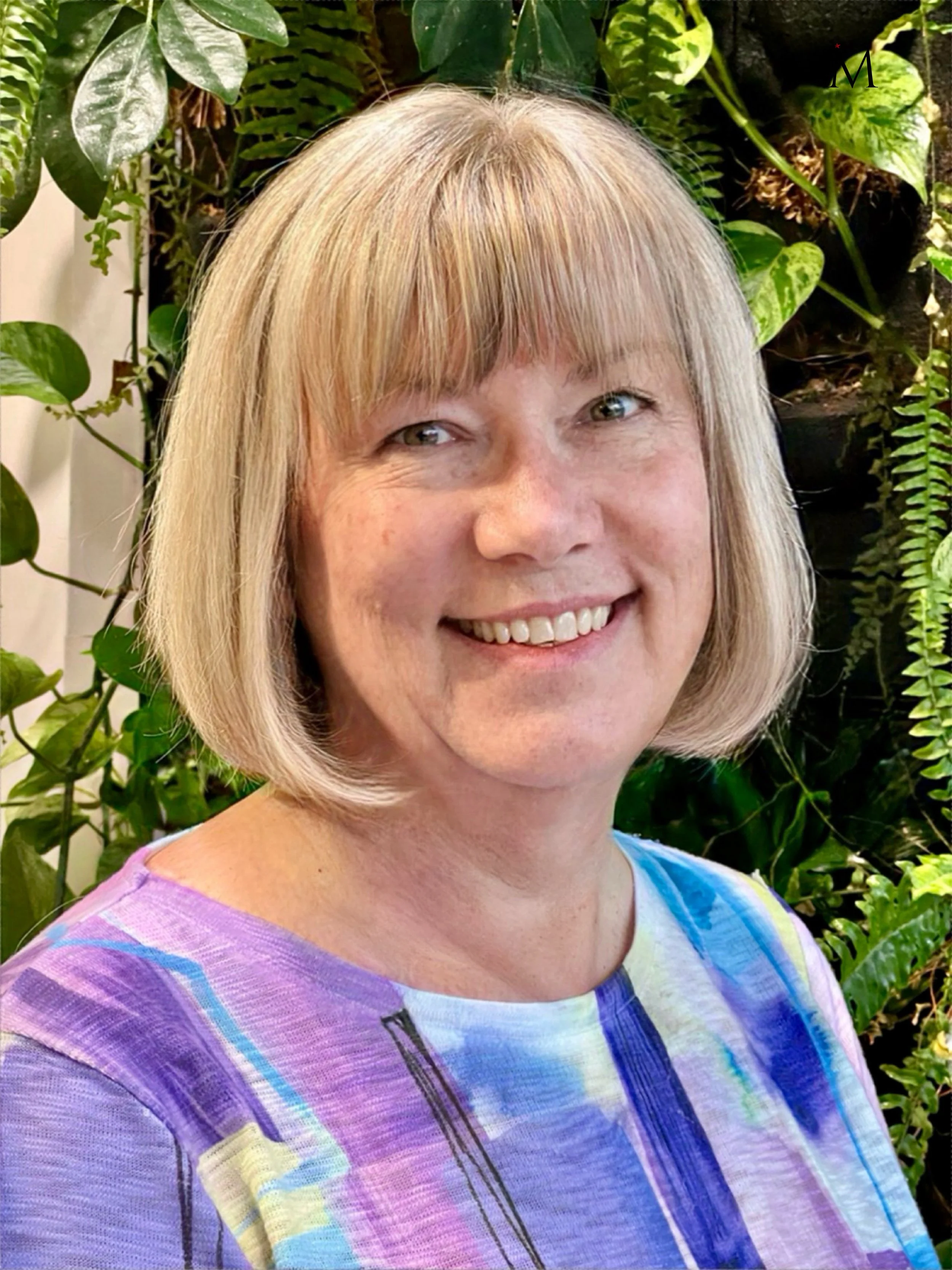 A woman with short blond hair smiling in front of a background with green plants.