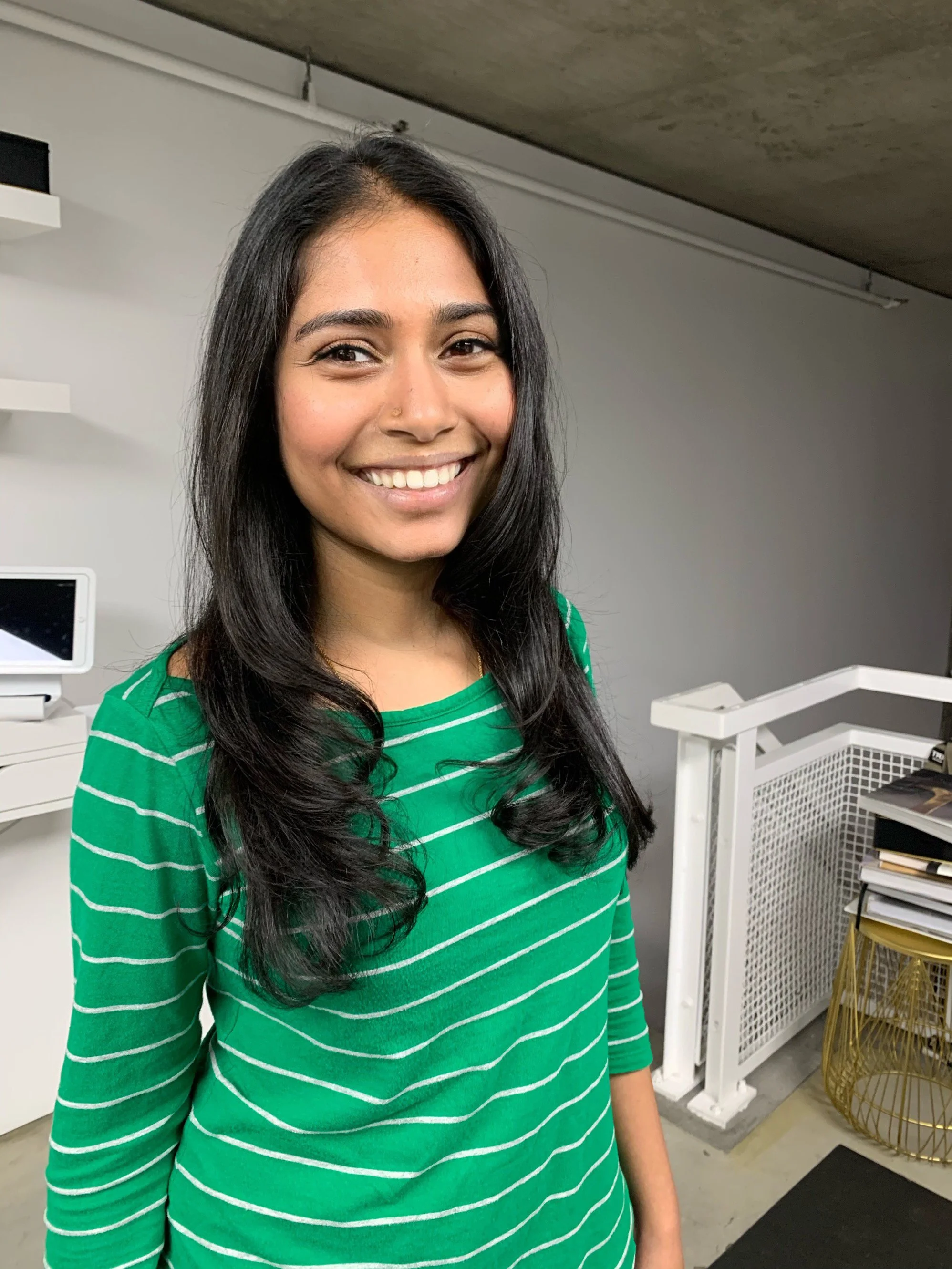 Young woman with long black hair and a nose piercing, smiling, wearing a green striped long-sleeve shirt, standing in an indoor setting with white walls and shelves.