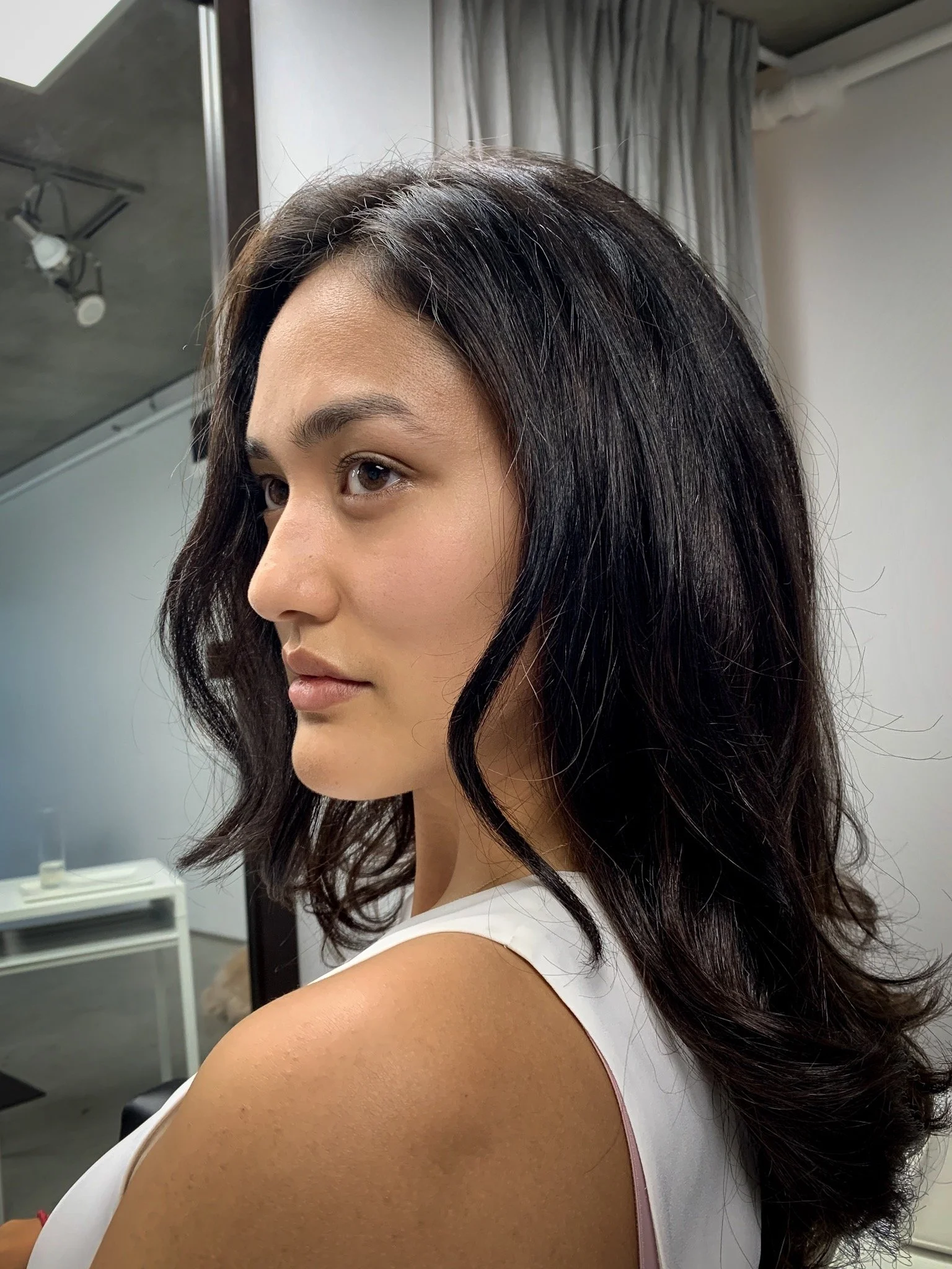 A young woman with shoulder-length wavy black hair, wearing a white sleeveless top, looking to her left in a room with neutral-colored curtains and a gray wall.