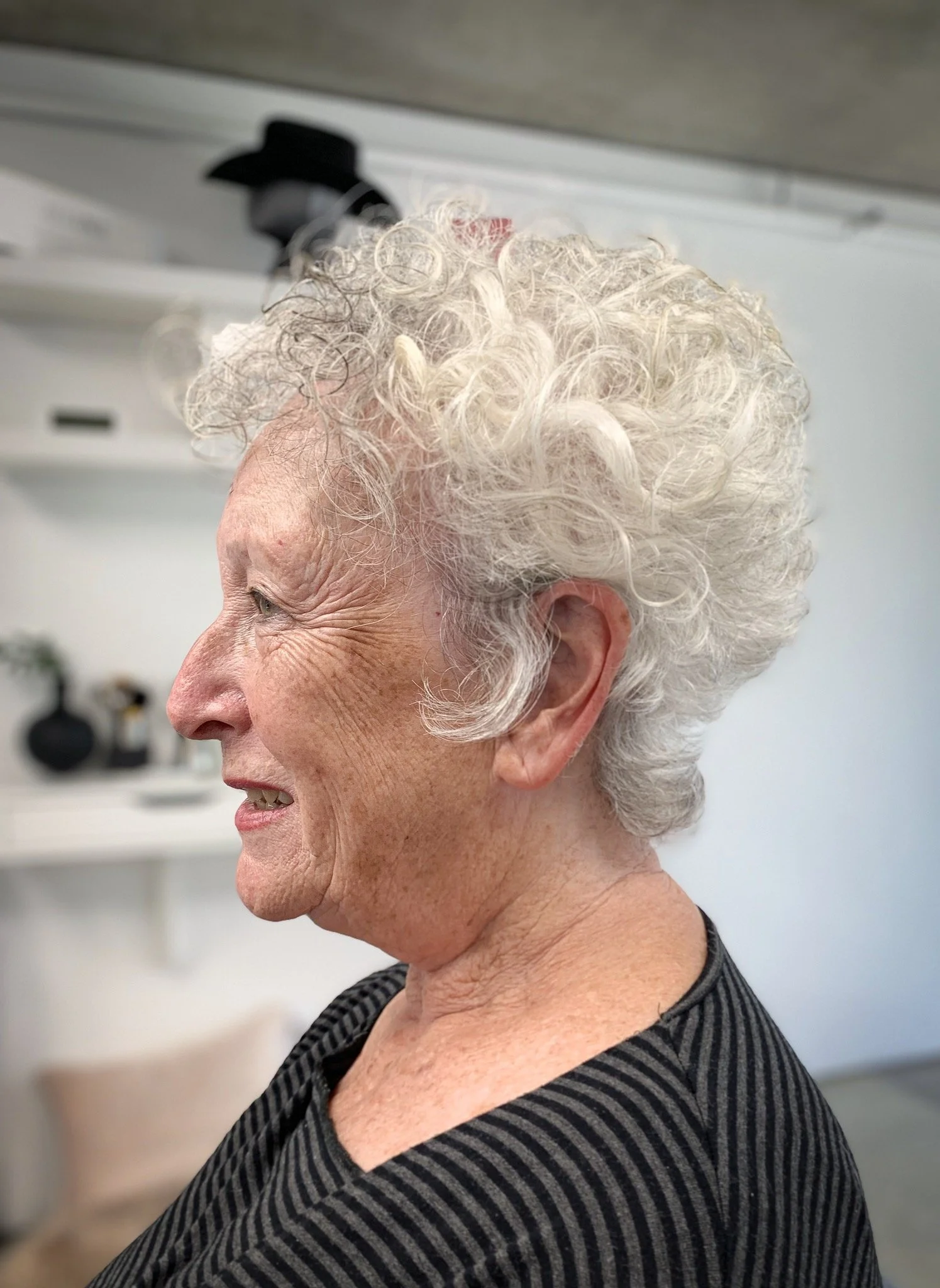 Profile of an elderly person with curly white hair, wearing a striped shirt, indoors.
