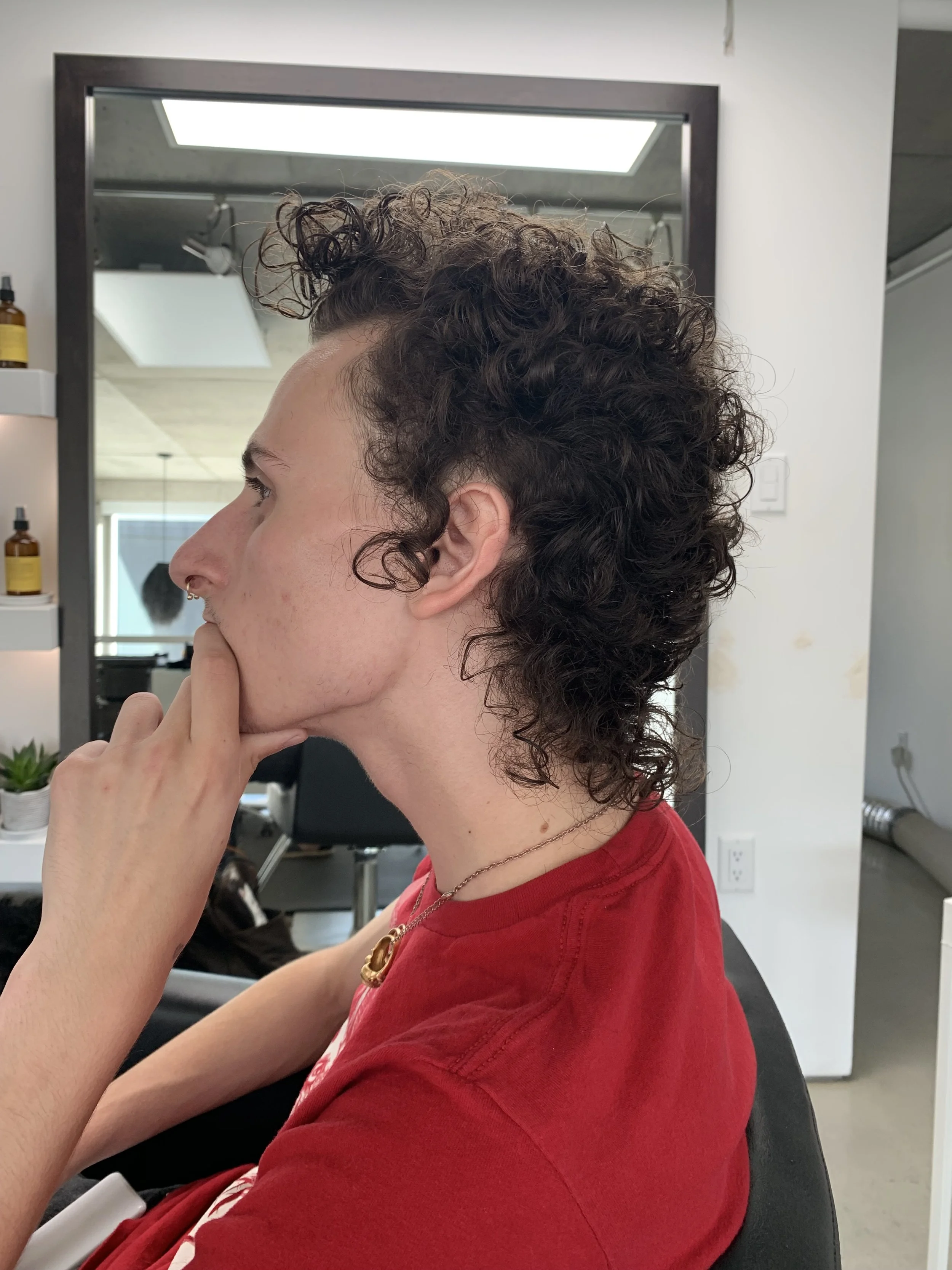 Person with curly hair sitting in salon, wearing red shirt, side profile view.