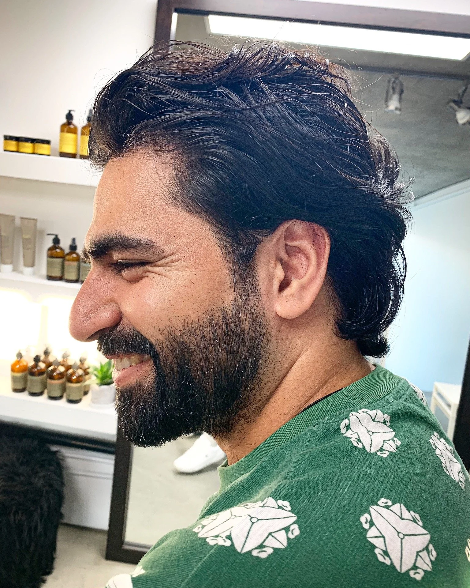 Side profile of a man with dark hair and a beard, smiling, wearing a green shirt with white geometric patterns, in a salon or barbershop setting.
