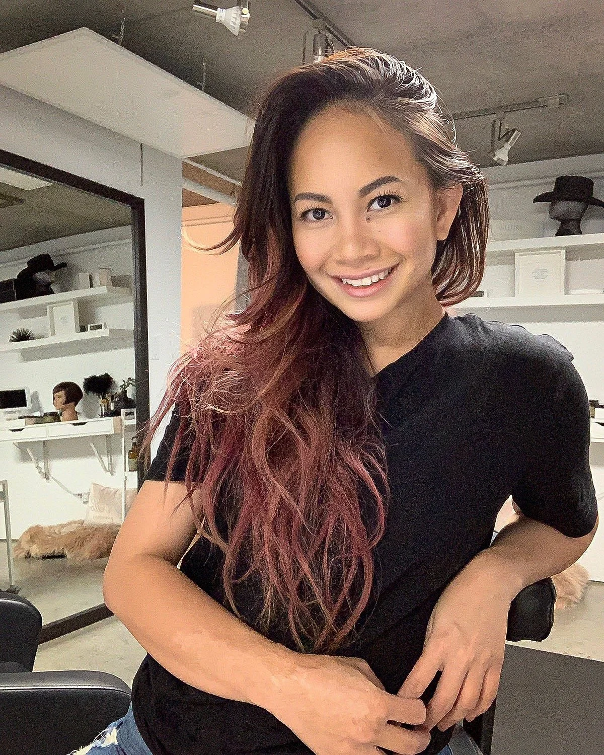 A young woman with long, wavy hair smiling at the camera in a modern indoor setting.