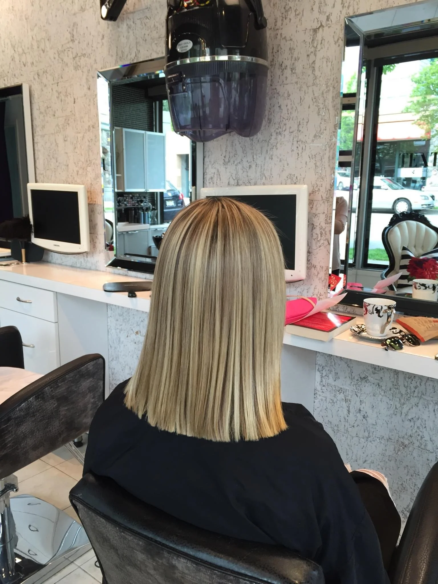 Woman with blonde, straight, shoulder-length hair sitting in a salon chair, facing away, in front of mirrors and salon equipment.