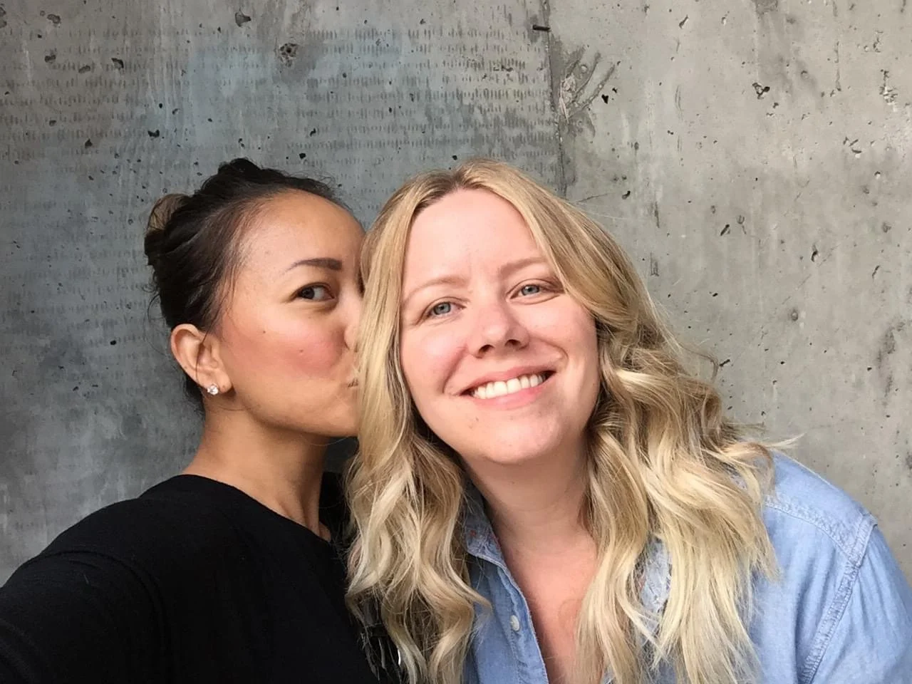 Two women, one with dark hair in a bun, giving a kiss on the cheek to the smiling woman with blonde wavy hair, against a textured concrete wall background.