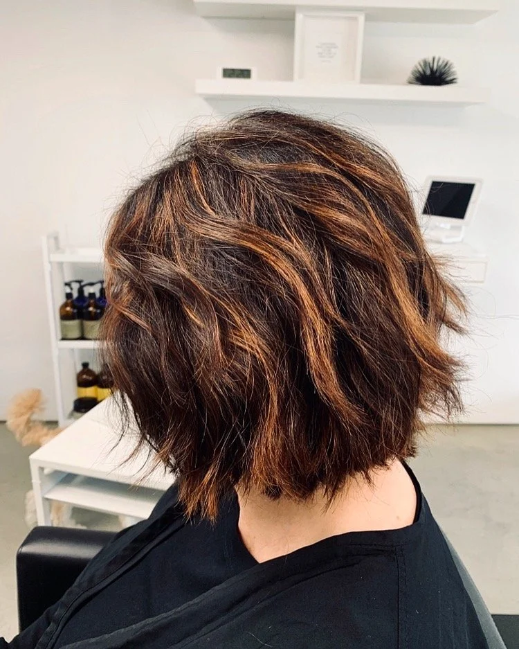 A woman with a short, wavy, layered haircut with brown and auburn highlights, sitting in a salon or spa with white shelves and bottles in the background.