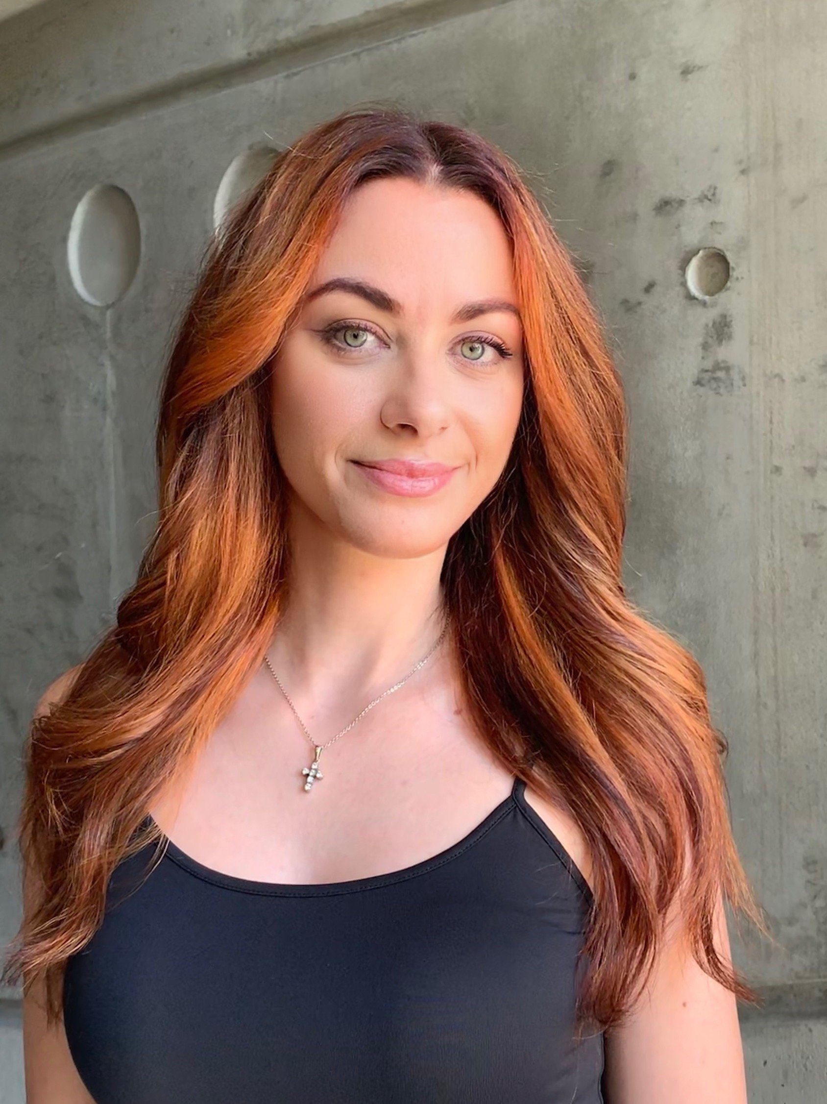 Woman with long red hair wearing a black top and a cross necklace against a concrete background.