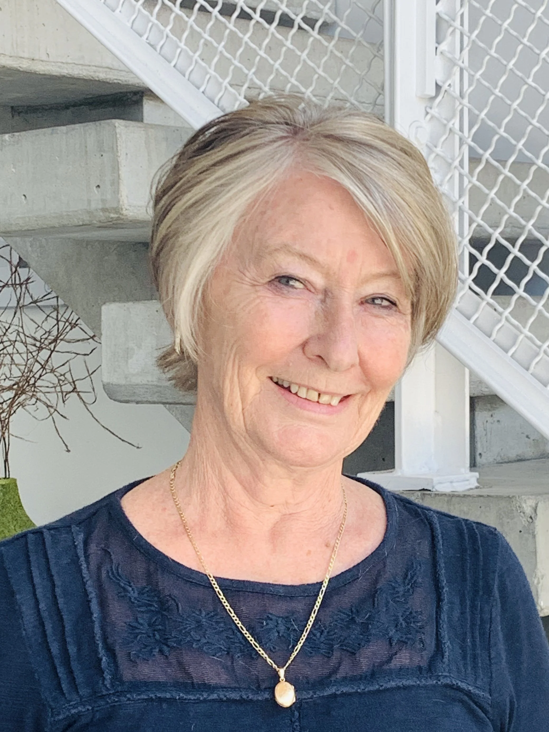 Elderly woman smiling, wearing a necklace and a dark blue top, standing in front of a stairway with a white railing.