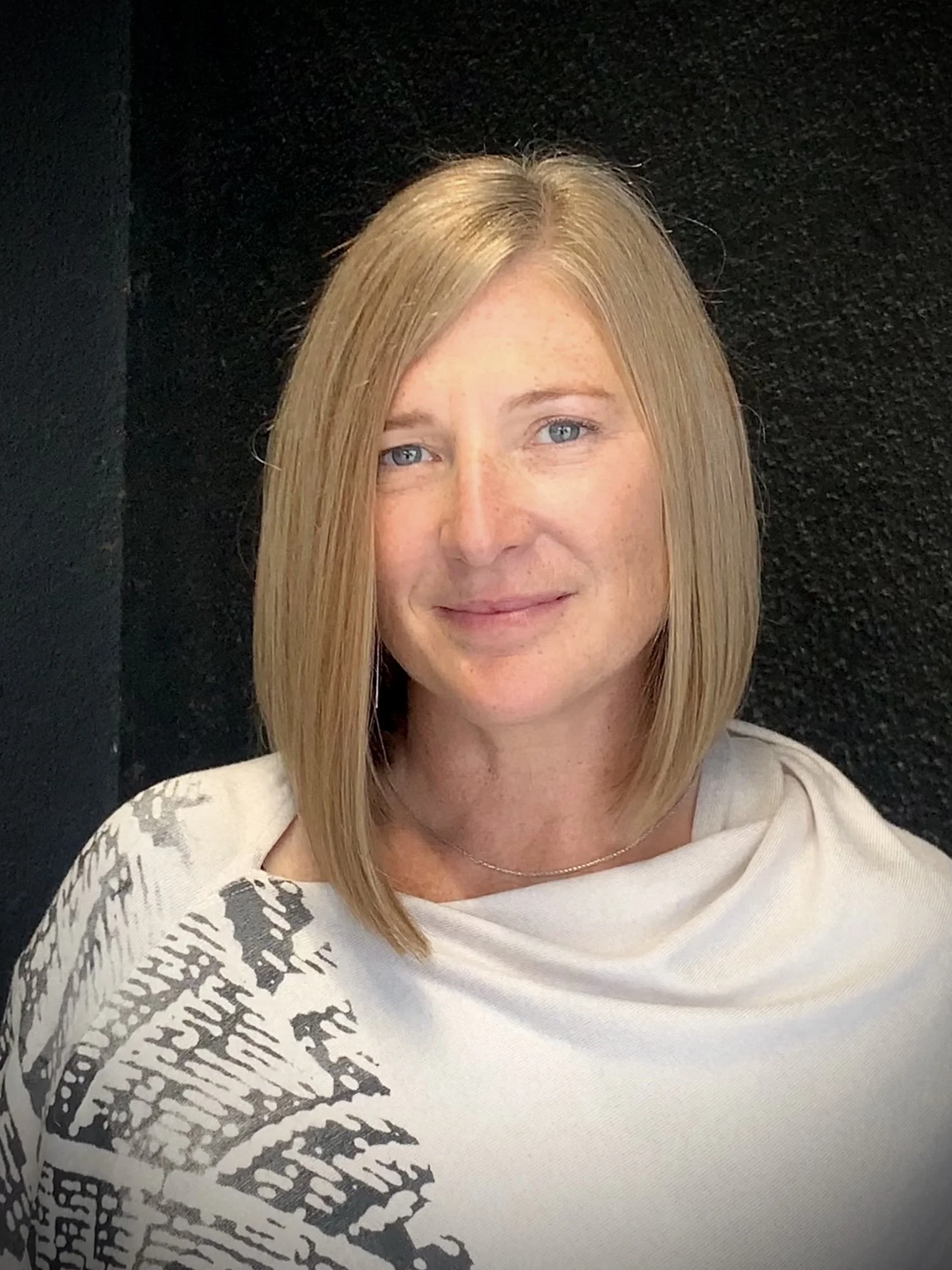 Woman with shoulder-length blonde hair and a patterned shawl, standing against a dark background.
