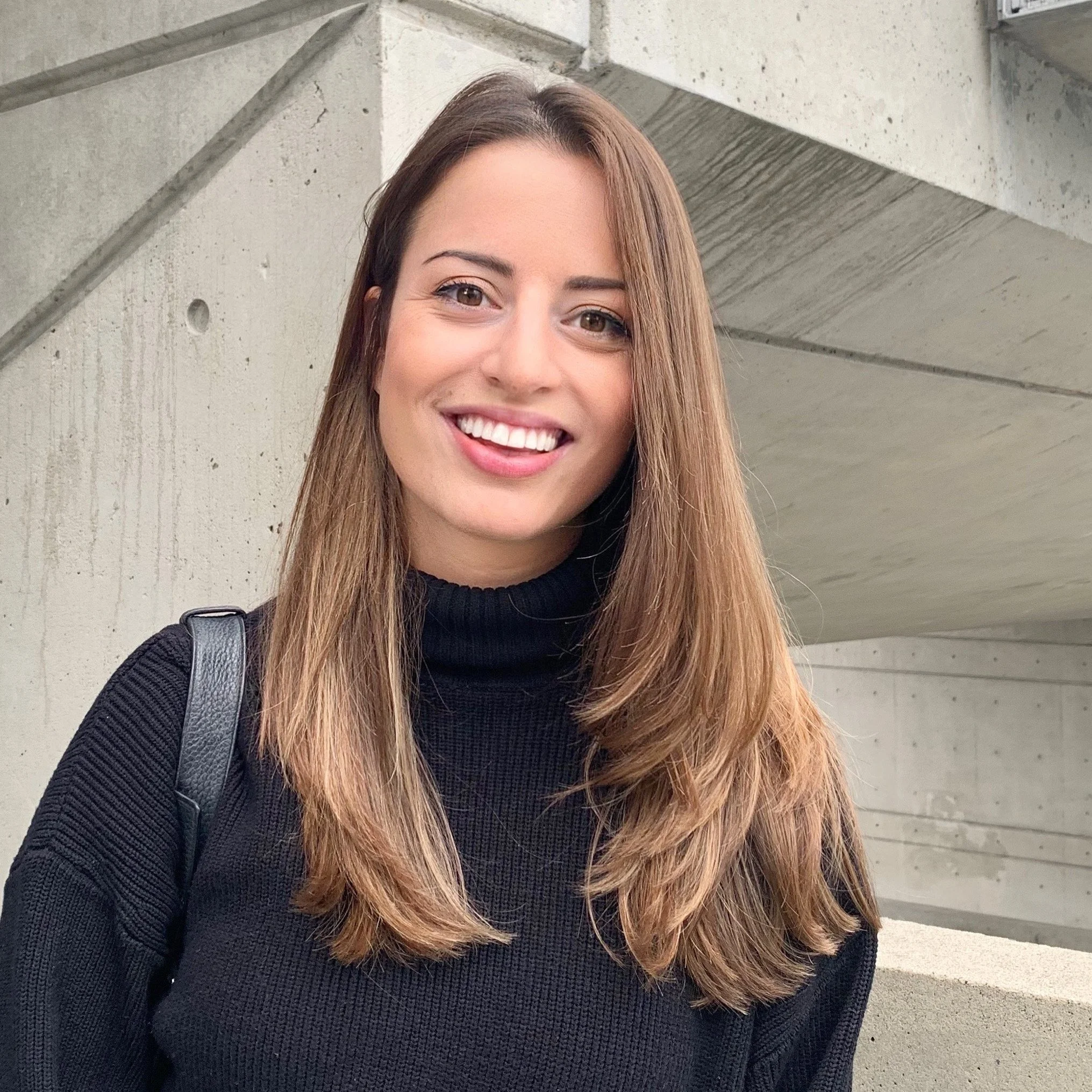 A young woman with long blonde hair, smiling, wearing a black turtleneck sweater, standing outdoors in front of a concrete structure.