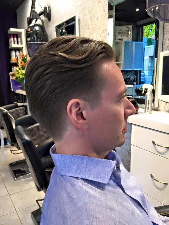 A man with styled, slicked-back hair sitting in a barber or Salon chair in a barbershop or salon, with various hair and beauty products and equipment visible in the background.