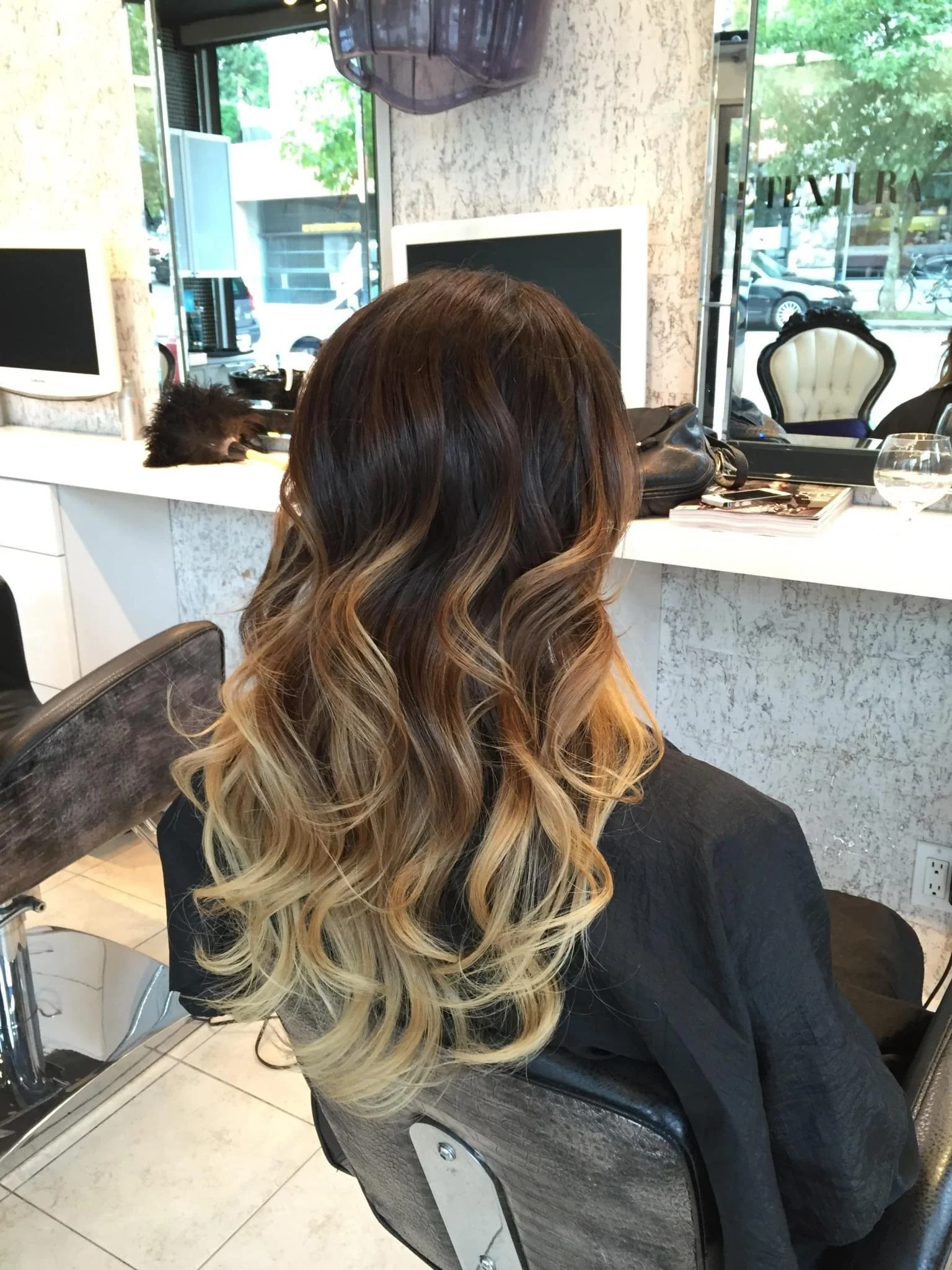 Back view of a woman with long wavy hair with brown roots and blonde tips, seated in a salon chair inside a hair salon.