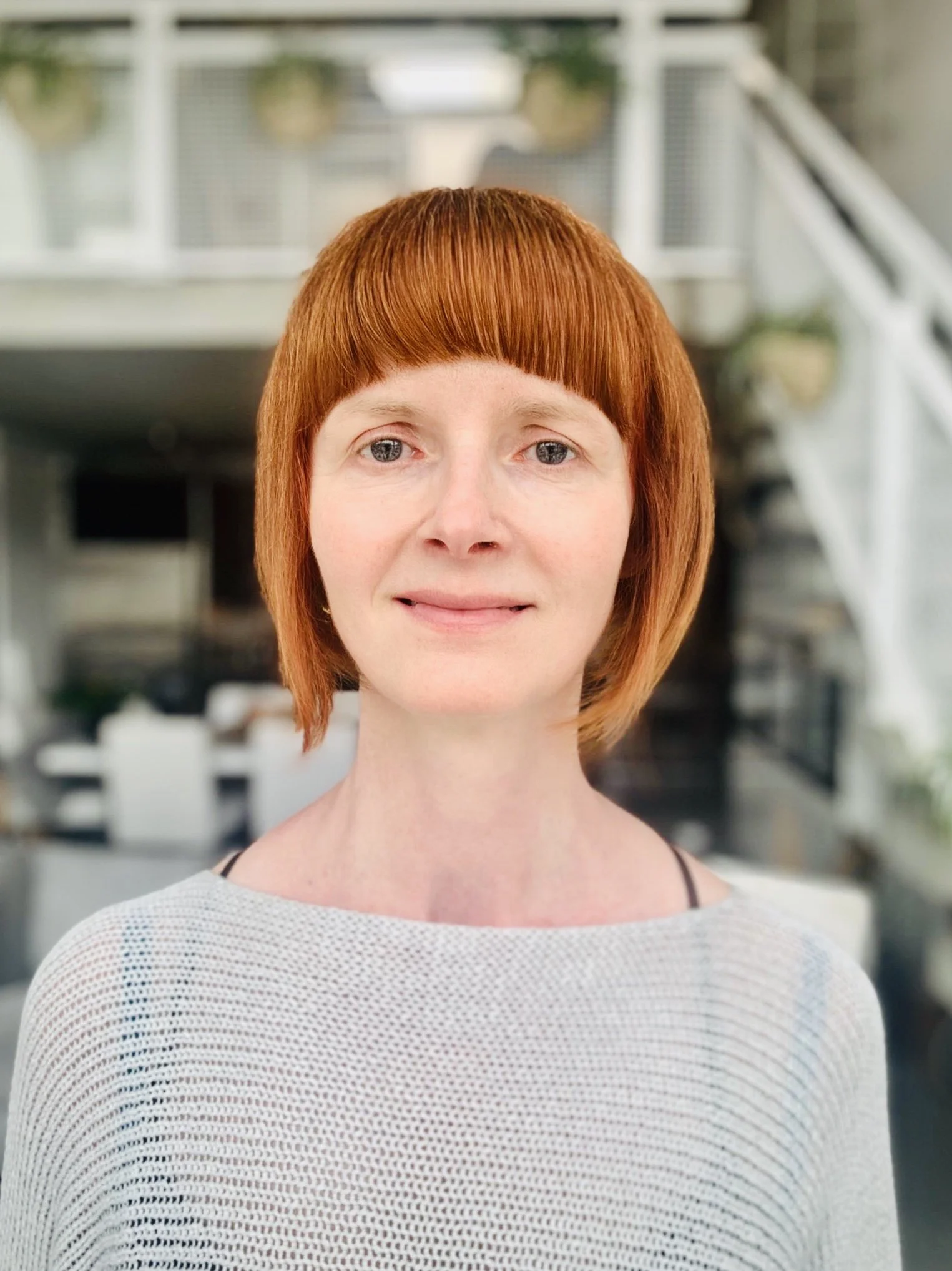 Person with short red hair and bangs, wearing a white top, standing indoors with a blurred background.