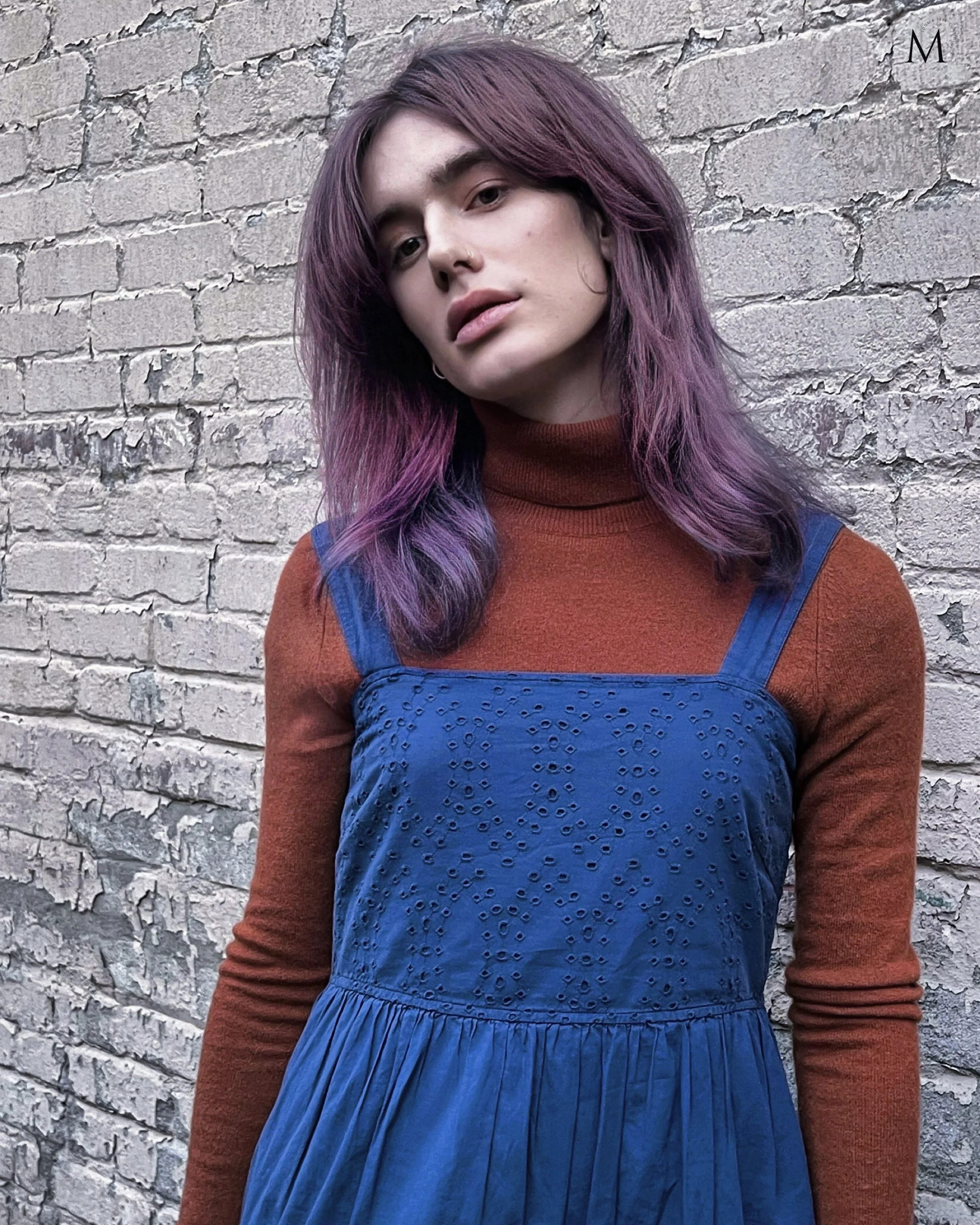 Person with long purple hair wearing a brown sweater and blue dress stands in front of a textured brick wall.