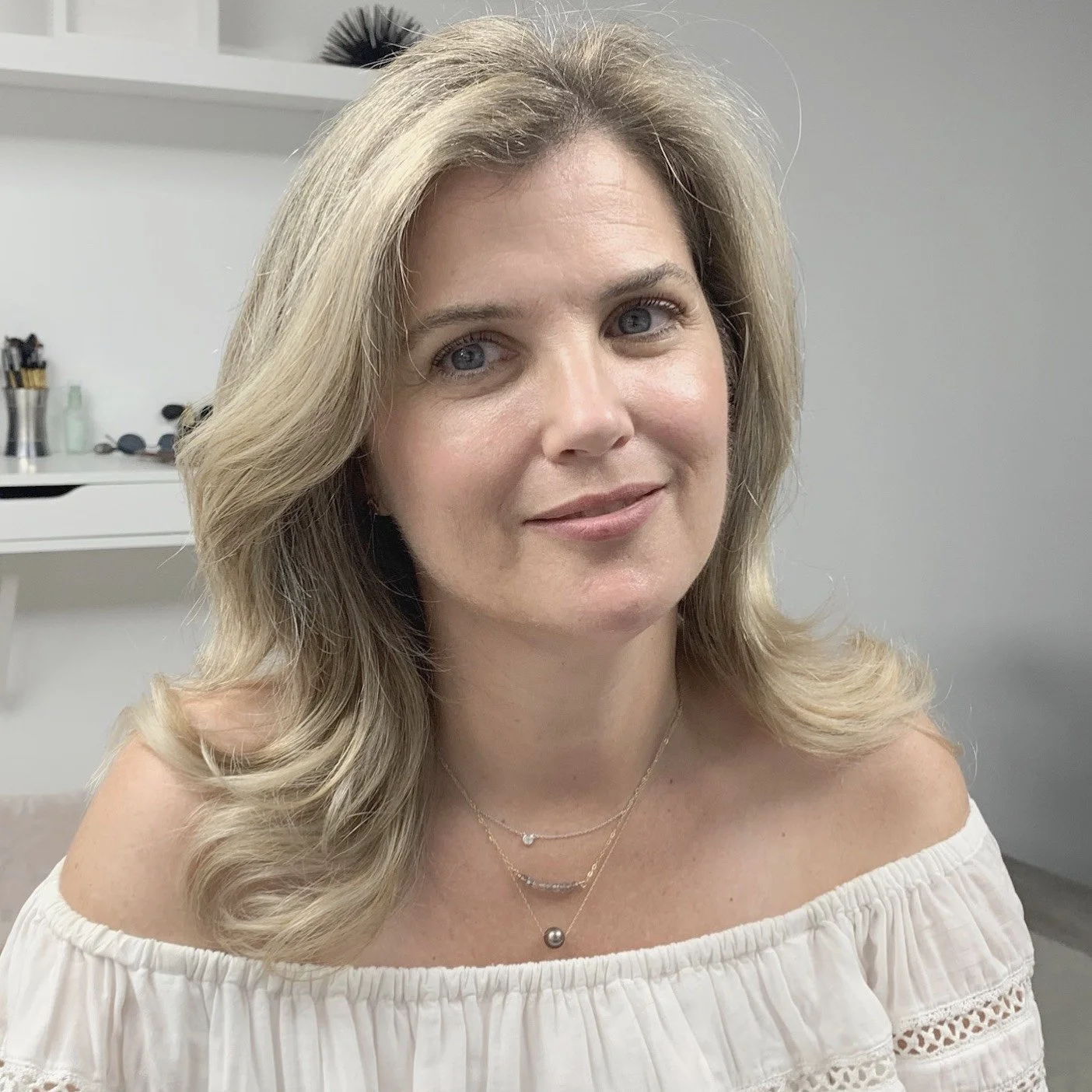 Woman with blonde hair smiling, wearing an off-shoulder white top, sitting indoors.