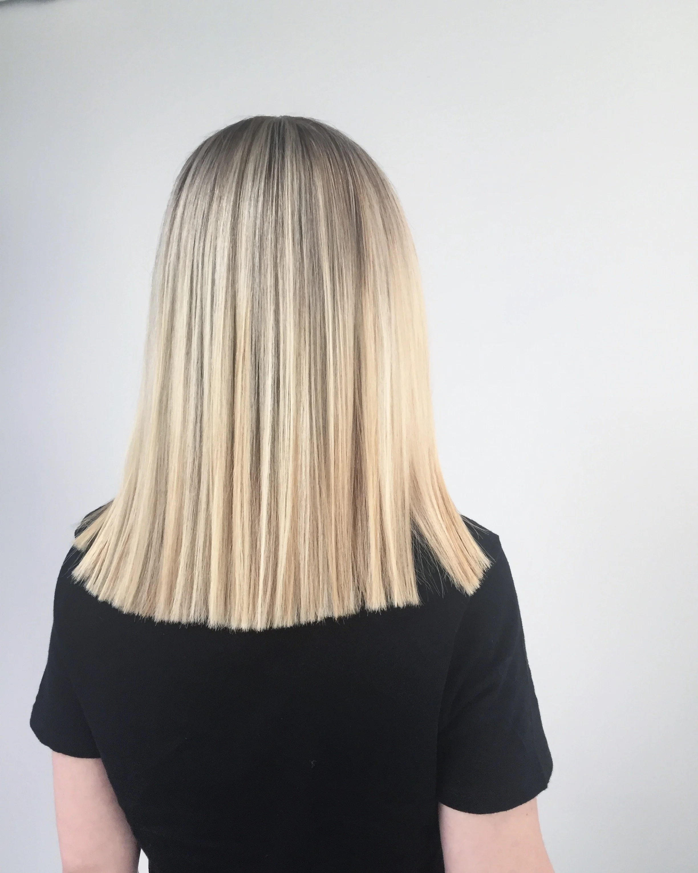 Back view of a person with straight, shoulder-length blonde hair, wearing a black shirt, standing against a white background.