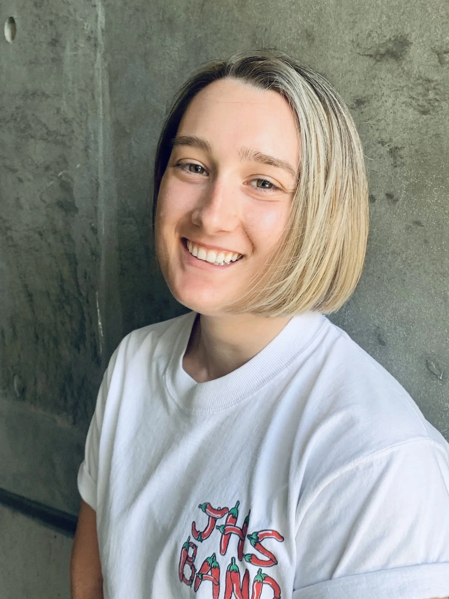 Person smiling in a white t-shirt with "JHS BAND" text, standing against a concrete wall.