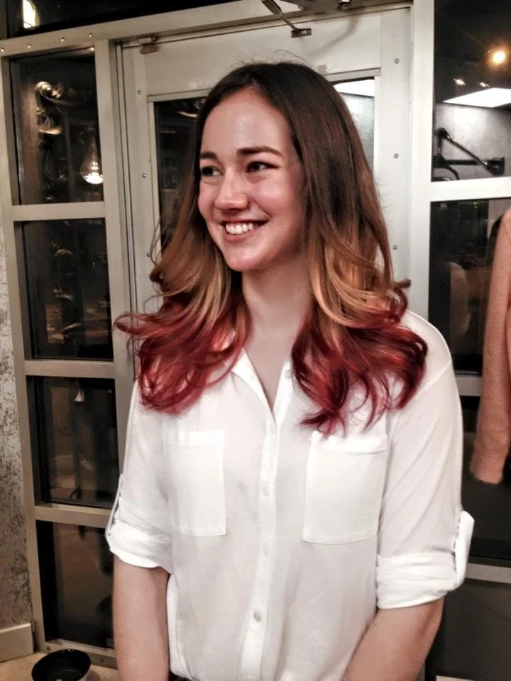 Young woman with long, wavy hair that is dark at the roots and fades to pink at the ends, smiling in an indoor setting with a metallic door and glass windows in the background.
