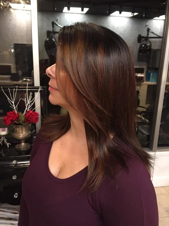 Side view of a woman with medium-length, shiny brown hair, wearing a dark purple top, standing indoors with a glass wall and a decorative vase with red flowers in the background.