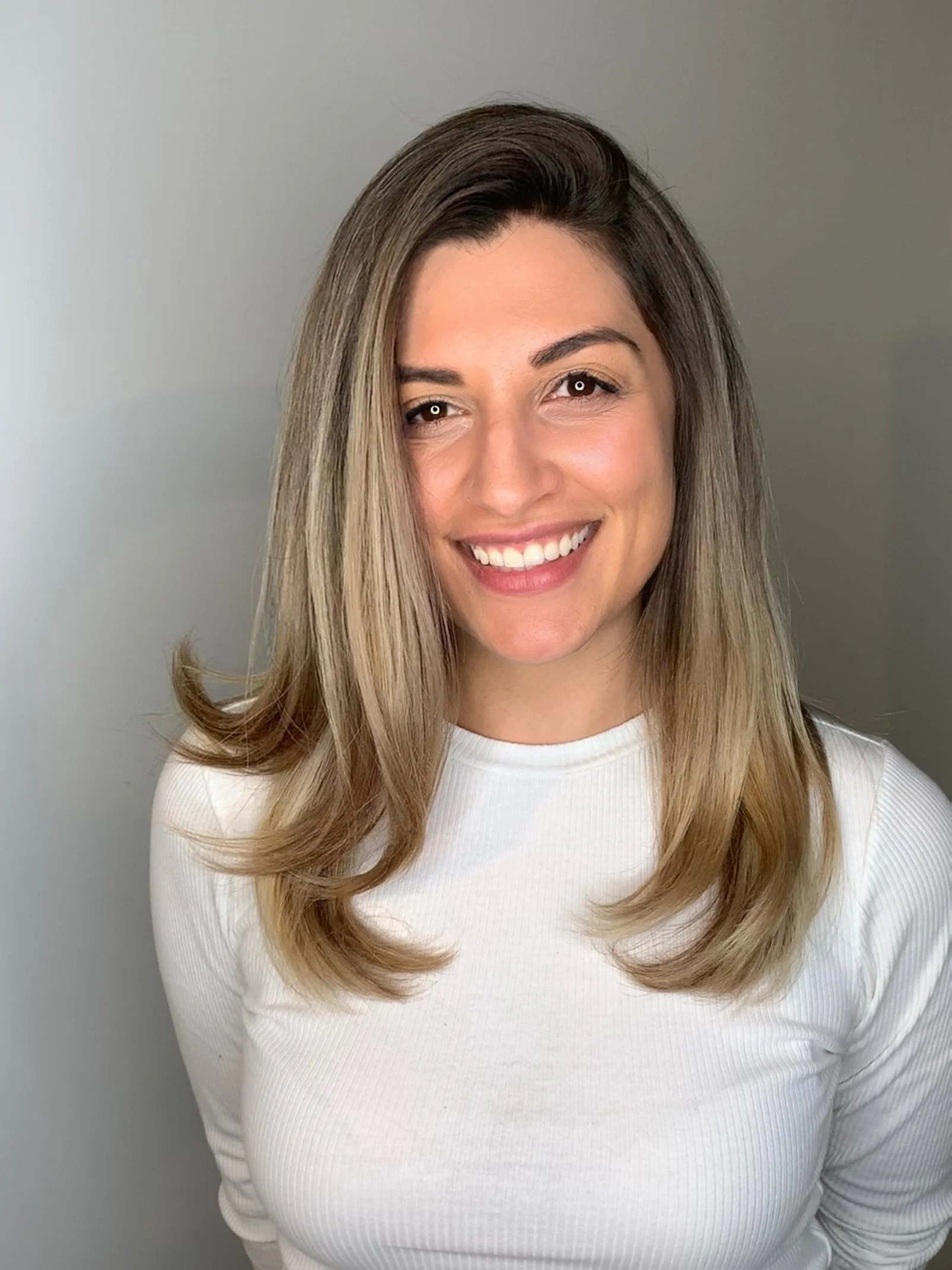 A woman with shoulder-length light brown hair, smiling, wearing a white ribbed shirt, standing against a plain gray background.