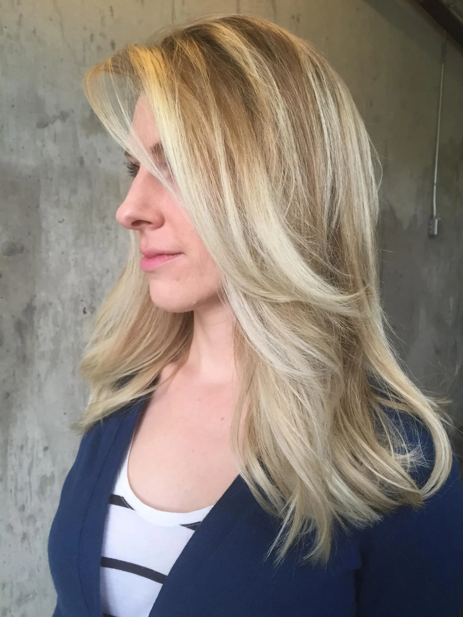A woman with long, wavy blonde hair wearing a dark blazer over a white top with black stripe details, standing against a concrete wall.