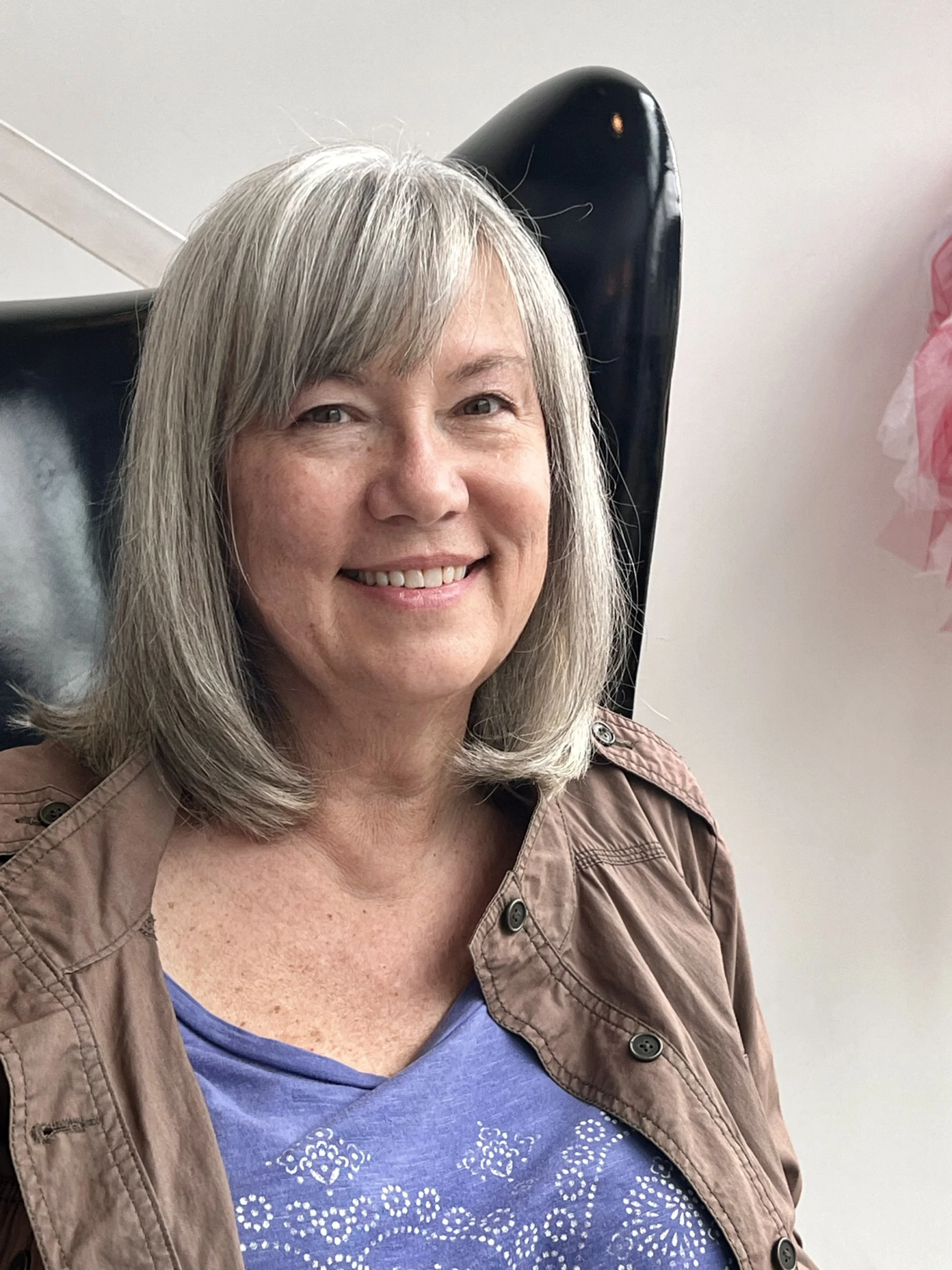 Smiling woman with gray hair wearing a brown jacket and blue shirt indoors.
