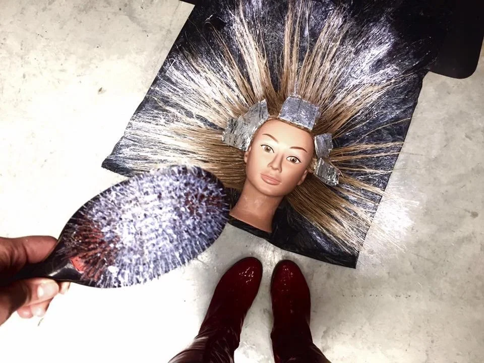Hairdressing mannequin head on a mat, with hair sectioned and prepared for coloring, surrounded by foils, with a hand holding a tint brush.