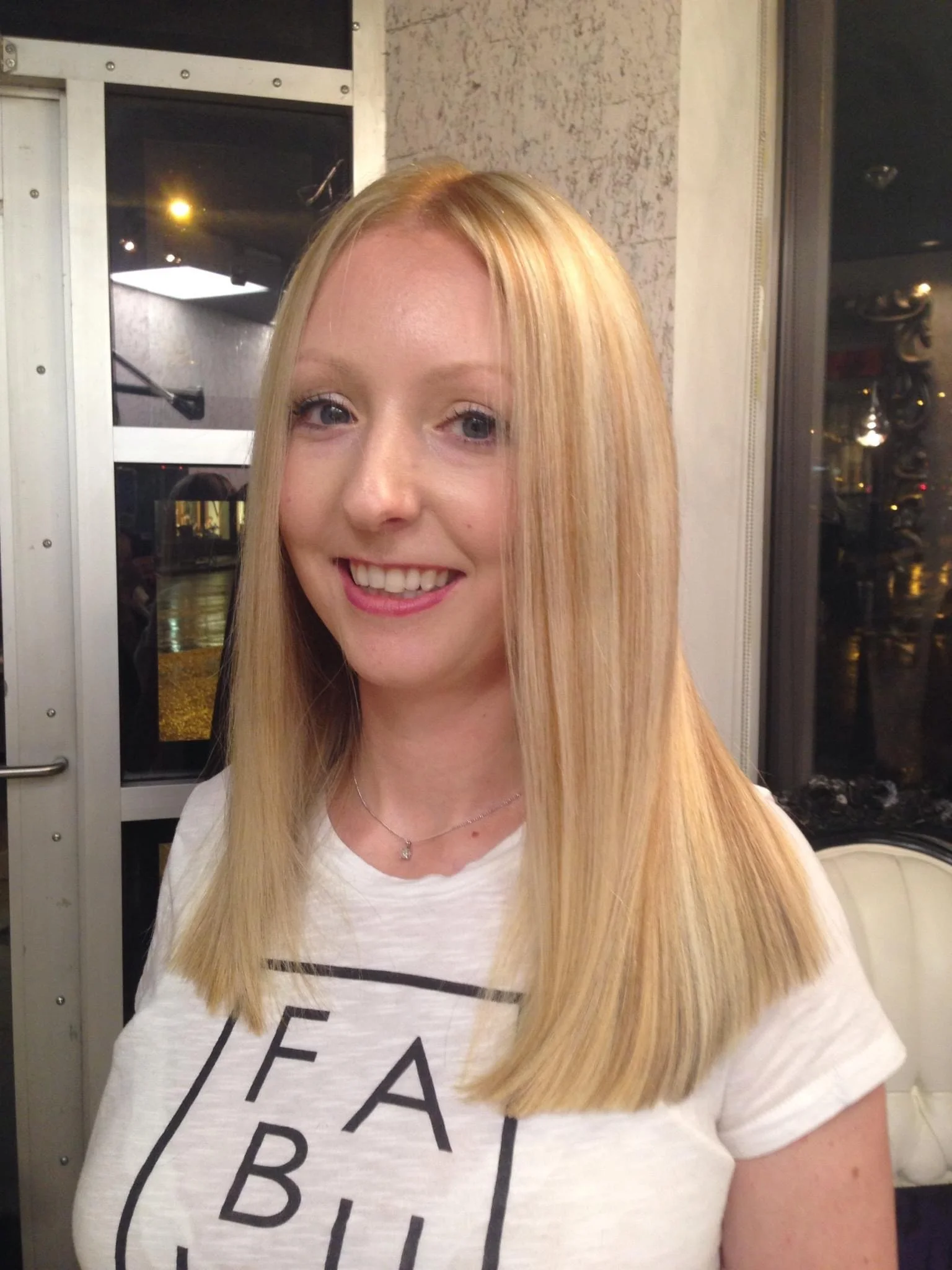 A woman with straight blonde hair wearing a white T-shirt with black text, smiling indoors at night.