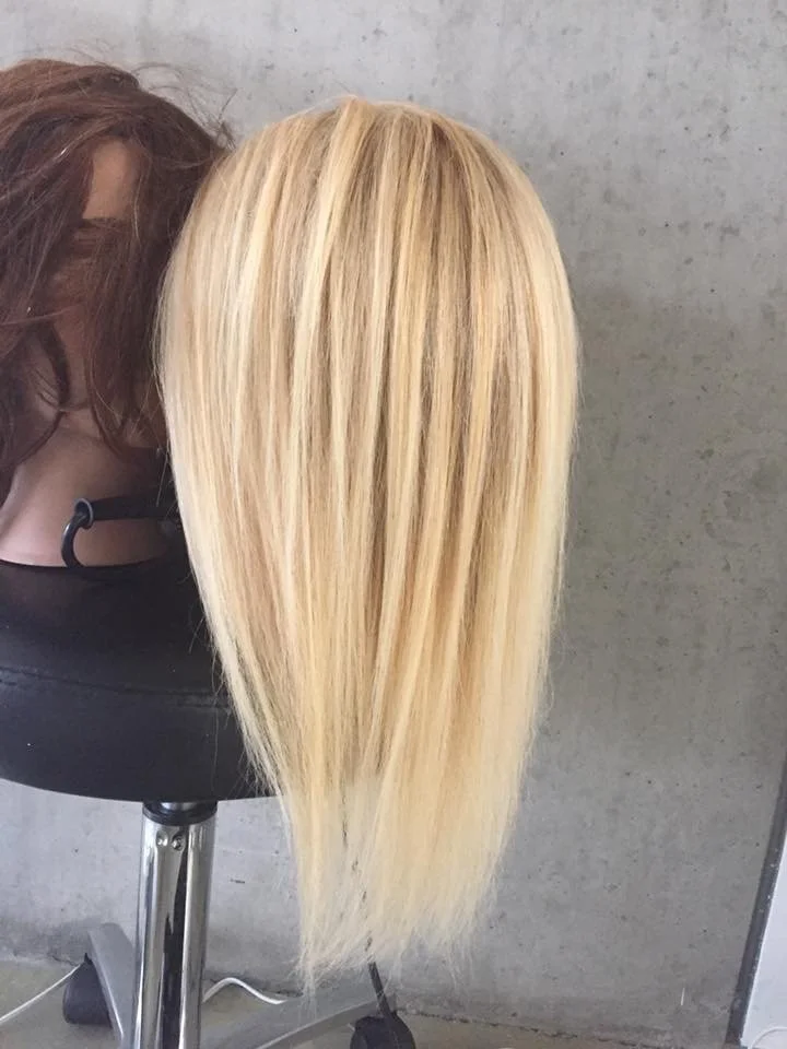 Blonde mannequin wig with straight hair on a black stand against a concrete wall.